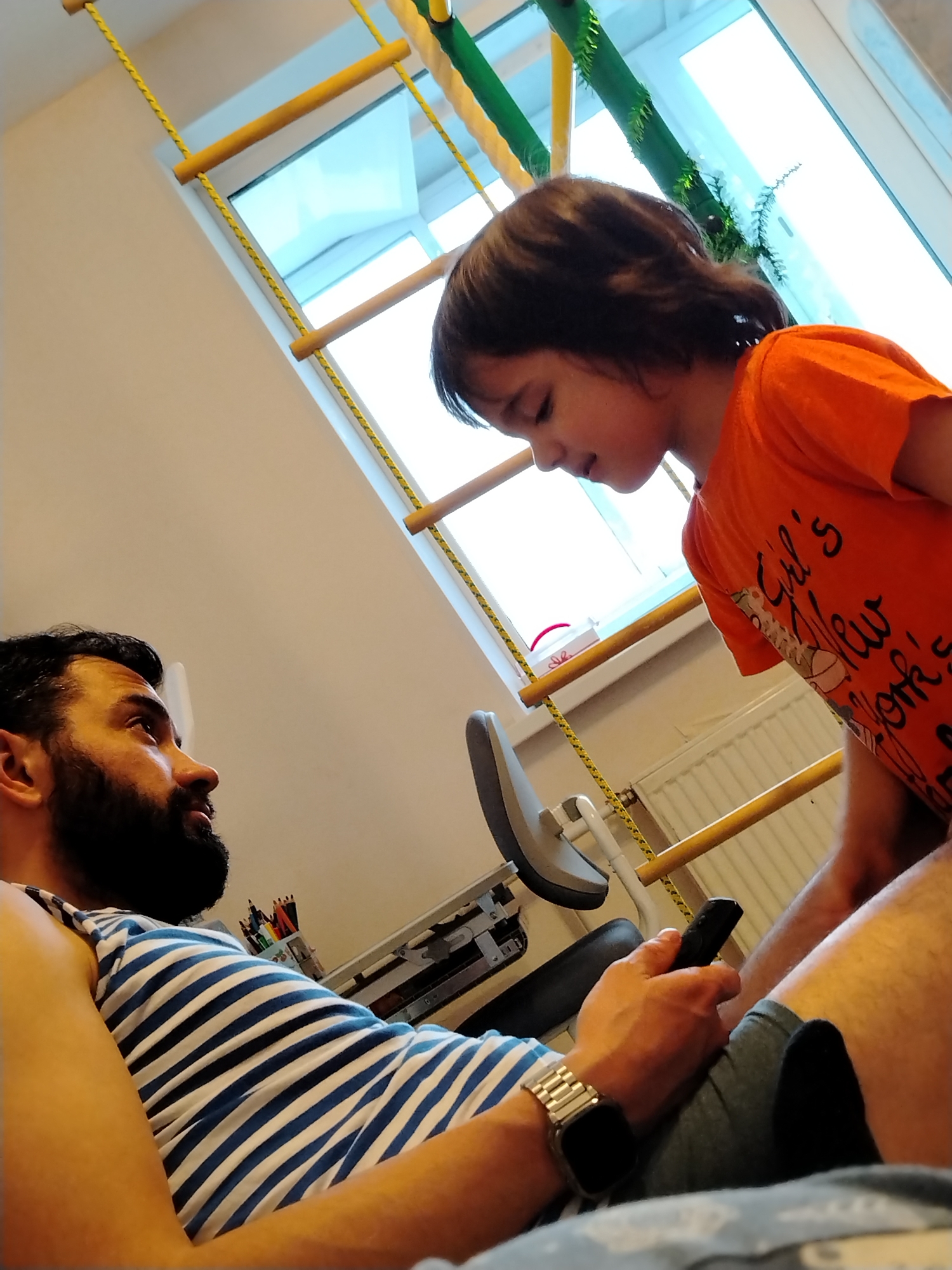 A medium shot captures a bearded man in a blue and white striped tank top reclining with a smartwatch on his left wrist and a black remote in his right hand, looking up at a young child wearing an orange t-shirt who stands above him. The backdrop includes a window with a yellow rope ladder structure and green decorative accents. Behind the man, a desk with colored pencils and pens is visible, along with a radiator. The location is Noviy, Russia.