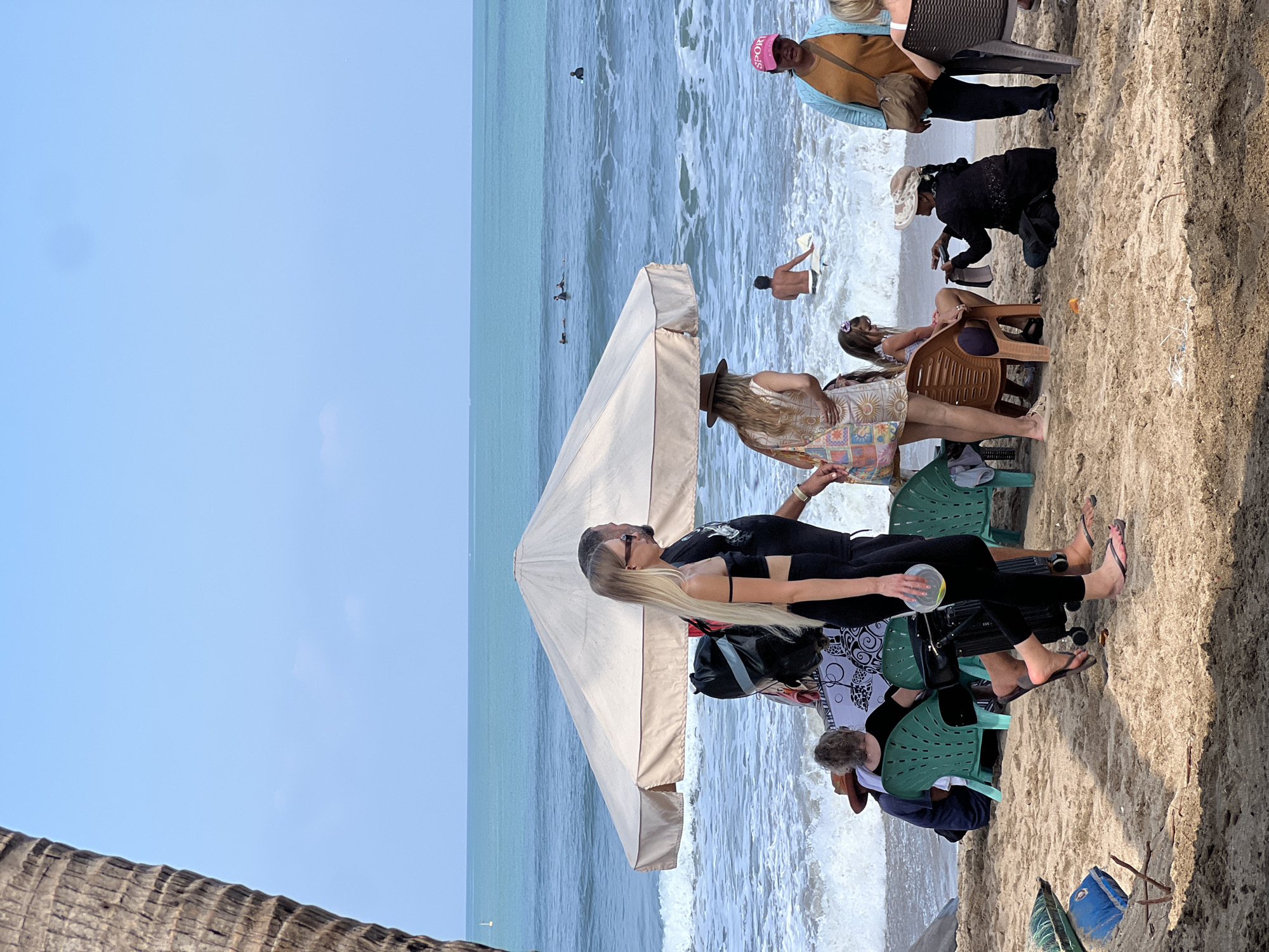 The image depicts a sunny day at a beach in Kuta, Indonesia, with the ocean in the background. Several people are present, some sitting on chairs, others standing, and some in the water.
In the foreground, a young woman with long blonde hair and sunglasses is walking to the right, wearing a black crop top and black leggings. Beside her, a man in a black t-shirt is also walking to the right. A large, cream-colored beach umbrella provides shade.
Further back, a woman with a patterned dress and a straw hat is standing. To her left, a young girl is seated on an orange chair. To the right, a woman in a blue cardigan and a pink hat is standing and looking towards the left. Another person is seated on a chair to the far right.
In the ocean, several people are surfing or swimming, with waves breaking on the shore. The sand on the beach is light brown. A palm tree trunk is visible on the far left edge of the image. The overall atmosphere appears relaxed and typical of a beach scene in a popular tourist destination.