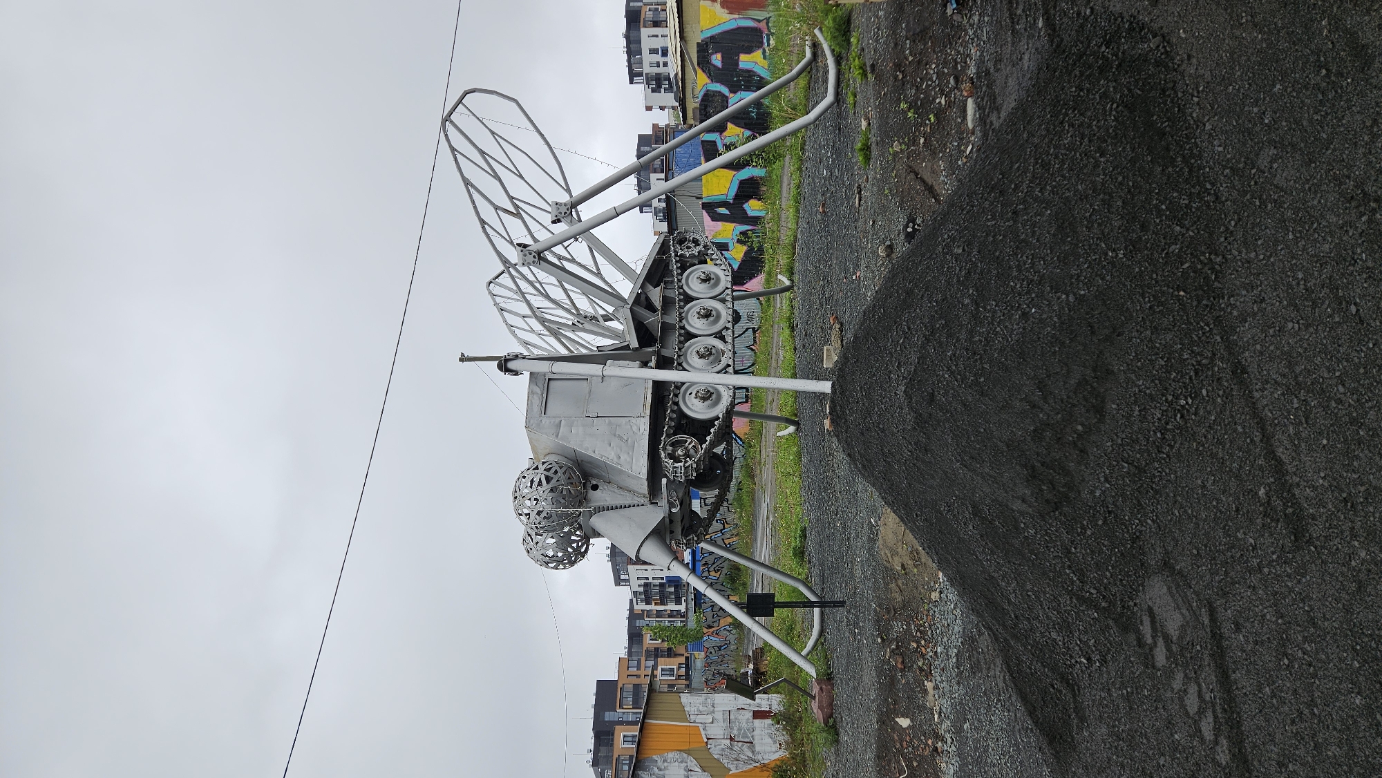 A metallic tank-like sculpture stands on a gravelly patch of land in an urban environment. The sculpture features tank treads, wheels, and what appears to be a metal cage structure on top. The front of the sculpture is adorned with two spherical, cage-like metal objects.
Behind the sculpture, there is a mix of graffiti-covered walls and modern apartment buildings under an overcast sky. A large pile of dark gravel or asphalt is located on the right side of the image. A utility wire is visible crossing the upper part of the frame.