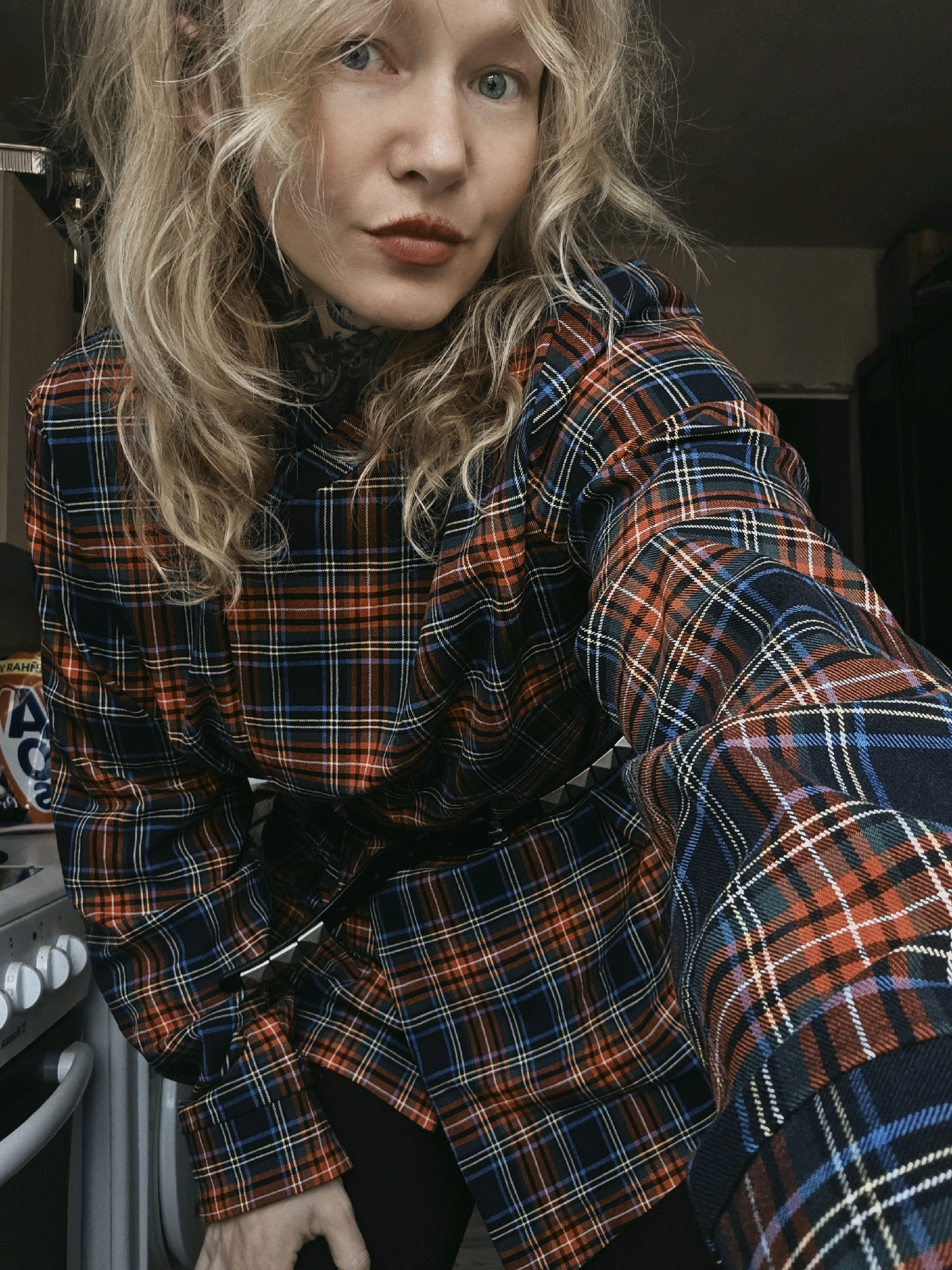 A woman with blonde, curly hair and blue eyes is pictured in a close-up shot. She is wearing a dark blue, red, and white plaid shirt. Her makeup includes red lipstick. She has tattoos on her neck. The background appears to be an indoor setting, possibly a kitchen, with a stove visible on the left side of the frame. The lighting is dim. The image quality is grainy. The location is identified as poselok Kornevo, Russia.