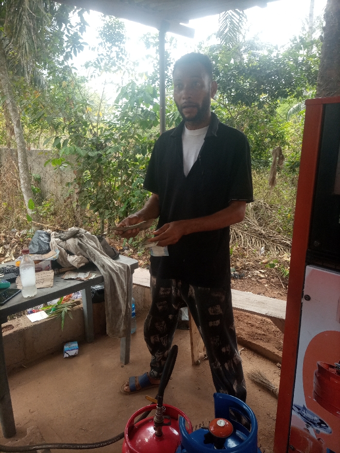 An adult male stands centrally within an outdoor LPG refilling station in Omicha Umu Awada, Nigeria. He possesses a dark beard and wears a black collared shirt over a white undershirt, dark patterned trousers, and blue sandals. The man faces the camera directly, holding bundles of Nigerian Naira currency in both hands, appearing to count them. To his left, a grey plastic table supports a clear plastic bottle with a handwritten label, a bundle of light-colored fabric, a calculator, a dark-colored smartphone, and other assorted papers and a black plastic bag. On the red-brown dirt ground, near the man's feet, are a red LPG cylinder and a blue LPG cylinder, both fitted with valves and connected by a black hose, indicative of a gas dispensing operation. A red-orange gas dispensing unit is partially visible on the far right. A small broom lies on the ground to the man's right. The background features dense green vegetation, including trees and bushes, a section of a concrete wall, and a chain-link fence. A rudimentary wooden bench is present behind the man. The scene is illuminated by natural daylight, casting soft shadows.