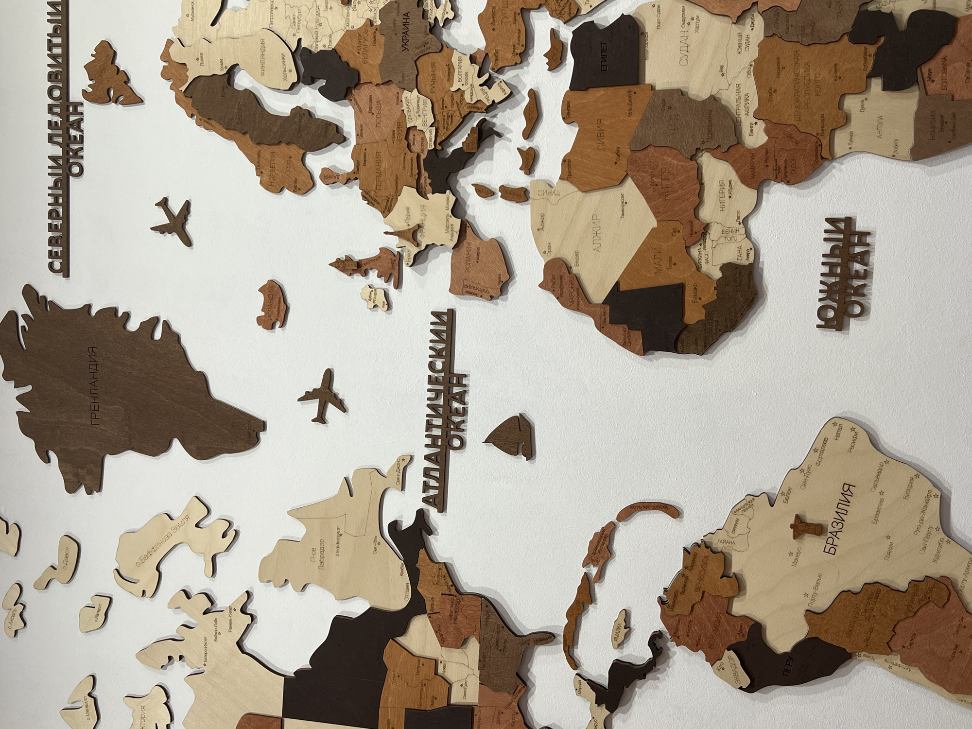 The image shows a wooden world map mounted on a white wall. The map is composed of individual wooden pieces representing continents, countries, and some islands. The pieces are different shades of brown, beige, and tan.
The continents visible in the image include North America, South America, Europe, Africa, and parts of Asia. Specific countries are labeled with names in Cyrillic alphabet, such as "Бразилия" (Brazil) and "Украина" (Ukraine). Major cities like "Париж" (Paris) and "Москва" (Moscow) are marked with a star symbol. Oceans are labeled with their names in Cyrillic alphabet, such as "Атлантический Океан" (Atlantic Ocean) and "Северный Ледовитый Океан" (Arctic Ocean).
There are also wooden airplane cutouts placed near Greenland and over the Atlantic Ocean, adding a decorative touch to the map. The background is a smooth, white wall. The map appears to be a decorative piece intended to provide geographic information in an aesthetically pleasing manner. There is nothing in the image that indicates it's specifically located in Yekaterinburg, Russia.
