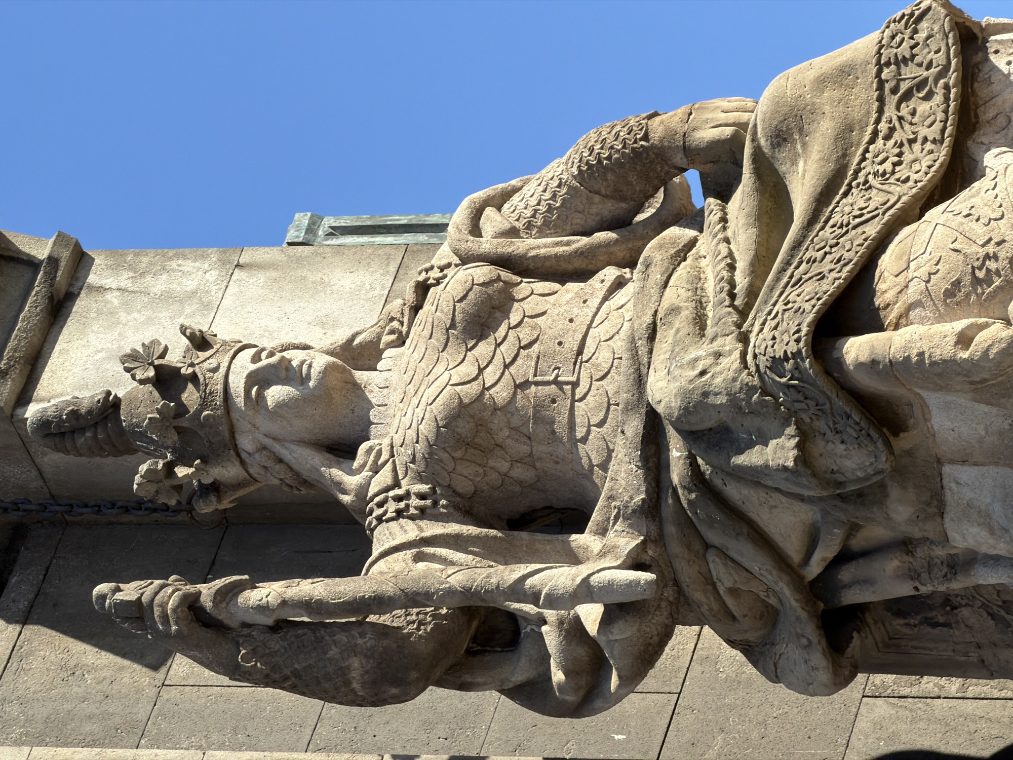 The image shows a close-up, low-angle, eye-level view of a seated stone statue of a regal female figure, likely a queen, against a clear blue sky. The statue is crafted from weathered stone and depicts the figure wearing a crown adorned with floral motifs and holding a staff or scepter in her left hand. Her body is covered in armor with a scale-like pattern, and she is seated on a draped throne. The detail of the carving is evident in the intricate patterns on her clothing and armor. The statue is located on the Arc de Triomf in Barcelona, Spain, which is a triumphal arch located at the northern end of the Passeig de Lluís Companys. The arch was built by Josep Fontserè as the main access gate for the 1888 Barcelona Universal Exposition.