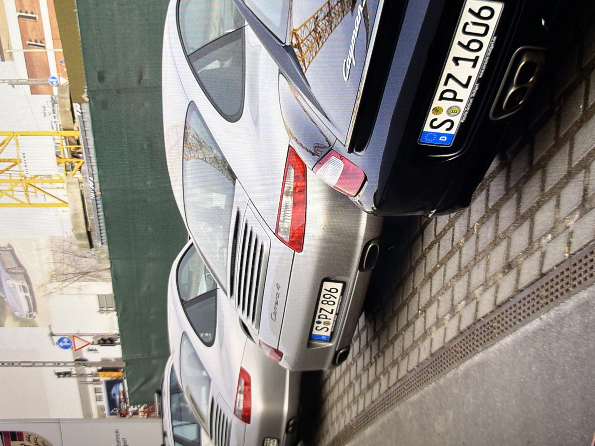 The image shows a row of parked Porsche cars. The cars are silver and black. The silver car in the foreground has a license plate that reads "S PZ 896". The black car in the foreground has a license plate that reads "S PZ 1606". The cars are parked on a brick pavement next to a drainage grate. In the background, there is a green fence, a yellow crane, and a brick building. The image does not show the city of Kyiv, Ukraine.
