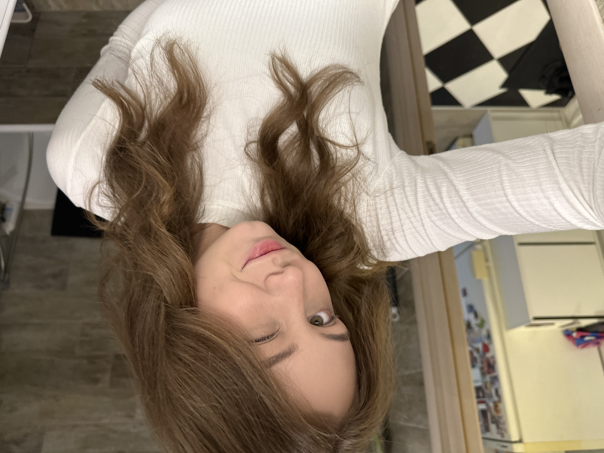 A woman with brown hair is taking a selfie indoors. She's wearing a white, long-sleeved shirt. The background shows part of a kitchen with a black and white checkered floor, cabinets, and appliances. The location is not definitively identifiable as Moscow, Russia based on the visible details.