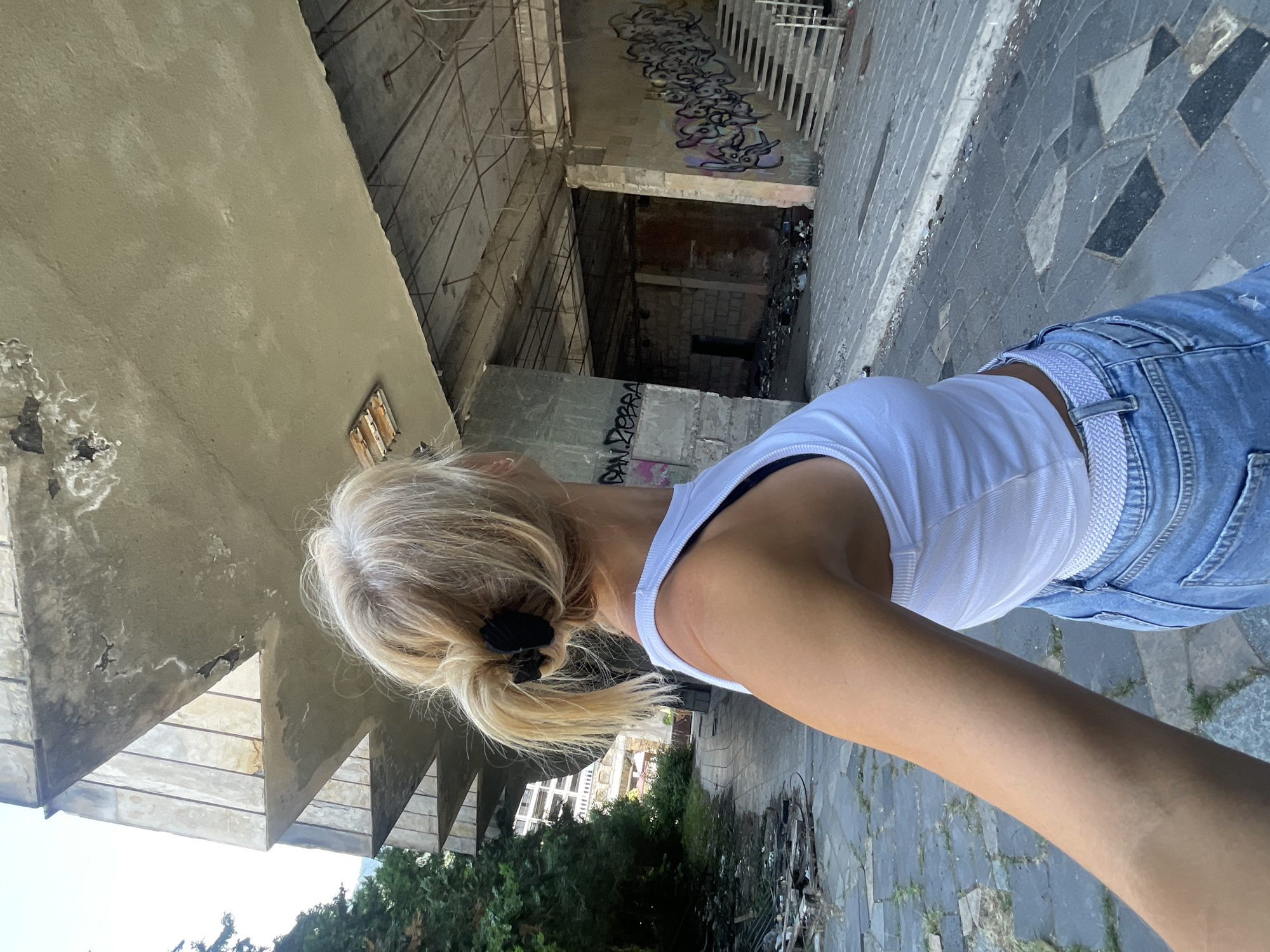 The image shows a blonde woman from the back, wearing a white crop top and blue jeans, standing in an abandoned, graffiti-covered building with concrete structures. She is looking up at the decaying architecture. The environment appears to be urban and neglected, with debris and overgrown vegetation visible. The specific location in Abaata, Abkhazia, is not discernible from the image alone.