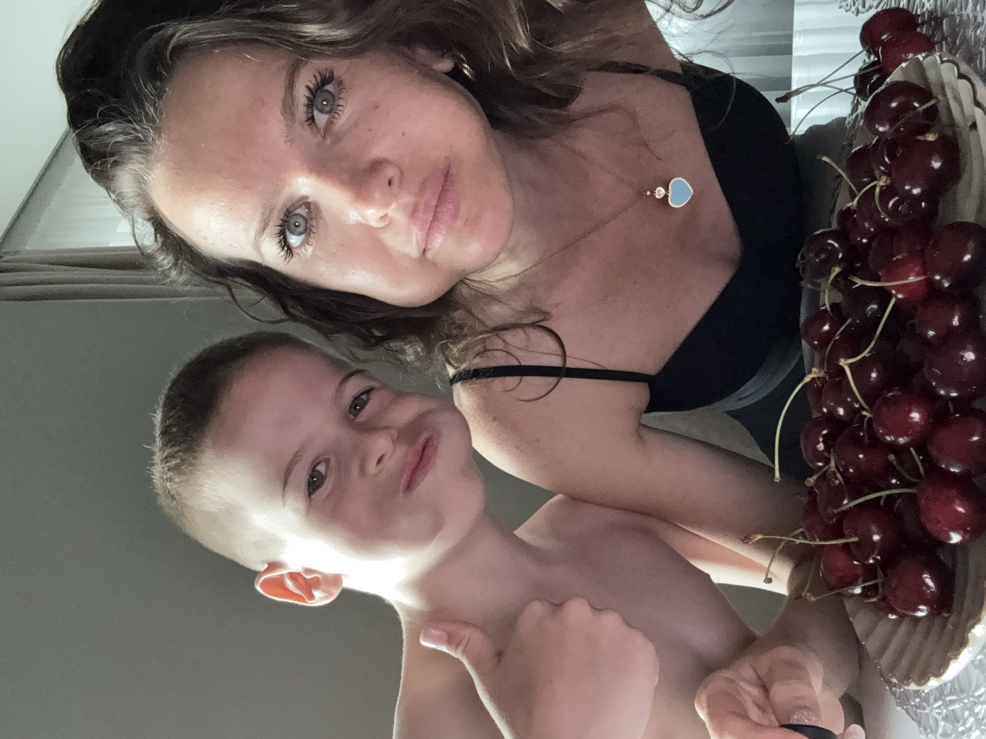 The media file is a selfie featuring a woman and a young boy. The woman has long, wavy brown hair and is wearing a black top. She is looking at the camera with bright, clear eyes and has a faint smile. Around her neck, she wears a delicate necklace with a heart-shaped pendant. Beside her, on the right side of the frame, is a bowl filled with a generous portion of fresh, dark red cherries.
The young boy, positioned in the foreground and slightly to the left, has short, light brown hair and is looking towards the camera with a playful expression, puckering his lips. He is shirtless and gives a thumbs-up gesture with his right hand. The image appears to be taken indoors, likely in a home environment given the close-up nature of the subjects and the presence of everyday objects like the bowl of cherries. The lighting suggests it could be daytime, possibly late afternoon or early evening. The overall mood is casual and intimate, capturing a moment between the two individuals. There is no visible text within the media file.