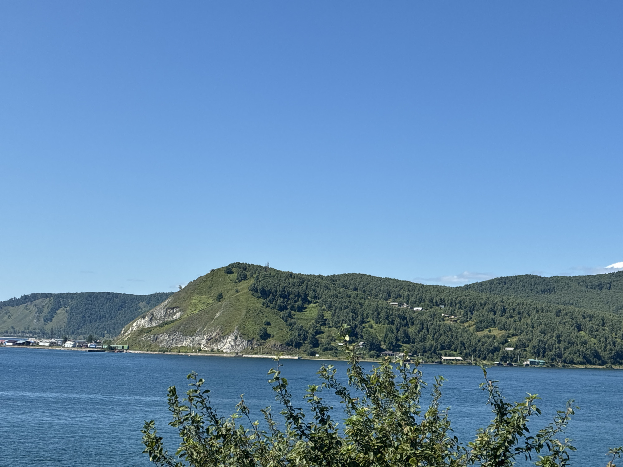 The image shows a wide, tranquil blue body of water, likely Lake Baikal, with a distant shoreline characterized by lush green, tree-covered hills and mountains. In the mid-ground, a prominent rocky hill juts out, its lighter-colored rock face contrasting with the dense vegetation that covers its slopes. Along the shoreline, a small settlement of buildings is visible, with some appearing to be clustered together, suggesting a village or small town. Further along the coast, more buildings are scattered amongst the trees. A few boats or barges are moored near the shore on the left side of the image. In the foreground, the branches of a leafy green bush or tree obscure the lower part of the scene. The sky is a clear, vibrant blue with no visible clouds. The overall impression is one of a serene, natural landscape with human habitation integrated into the environment. This scene is consistent with the general appearance of Listvyanka, Russia, a popular tourist village on the western shore of Lake Baikal.