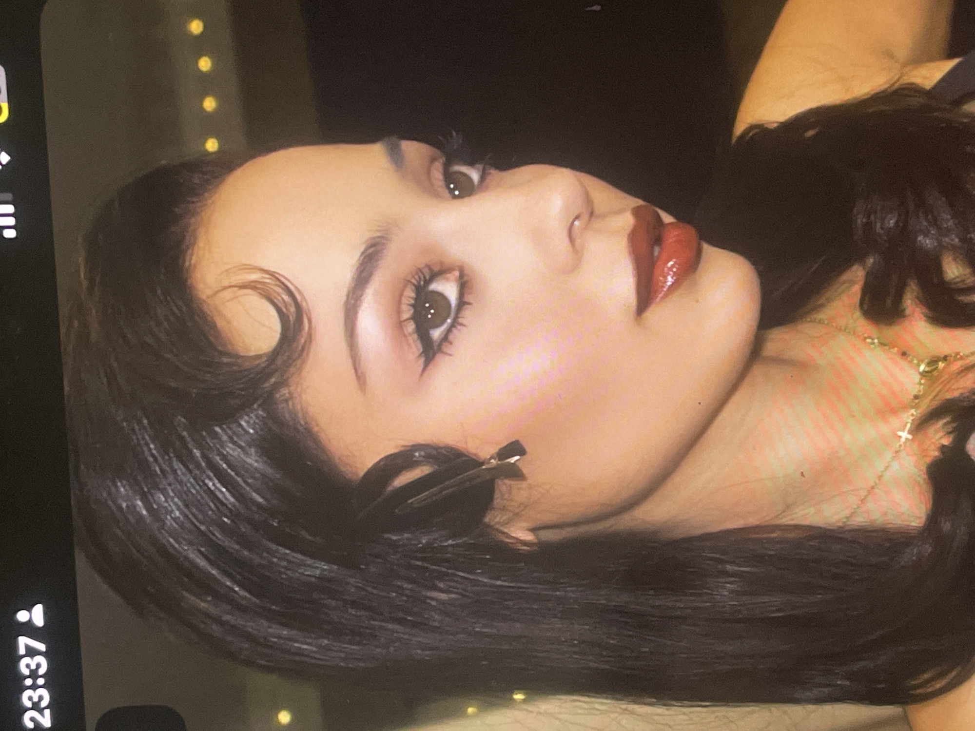 A close-up portrait of a young woman is depicted, with her head slightly tilted. She has long, dark hair, styled with a distinct finger wave curl near her forehead on the left side, and a black hair clip securing hair on the right. Her facial features are prominent, with defined eyebrows, dark eyes enhanced by winged eyeliner, and lips painted with glossy, dark red lipstick. Her expression is neutral, gazing upwards and to the side.
She wears a gold-colored necklace featuring a small cross pendant. The background is dark and out of focus, adorned with several blurred yellow circular lights, suggesting an indoor setting with ambient lighting. A digital overlay on the left side of the image displays "23:37," indicating the time of day is late evening. The image captures a stylish and contemplative moment. This portrait was taken in Poltava, Ukraine.