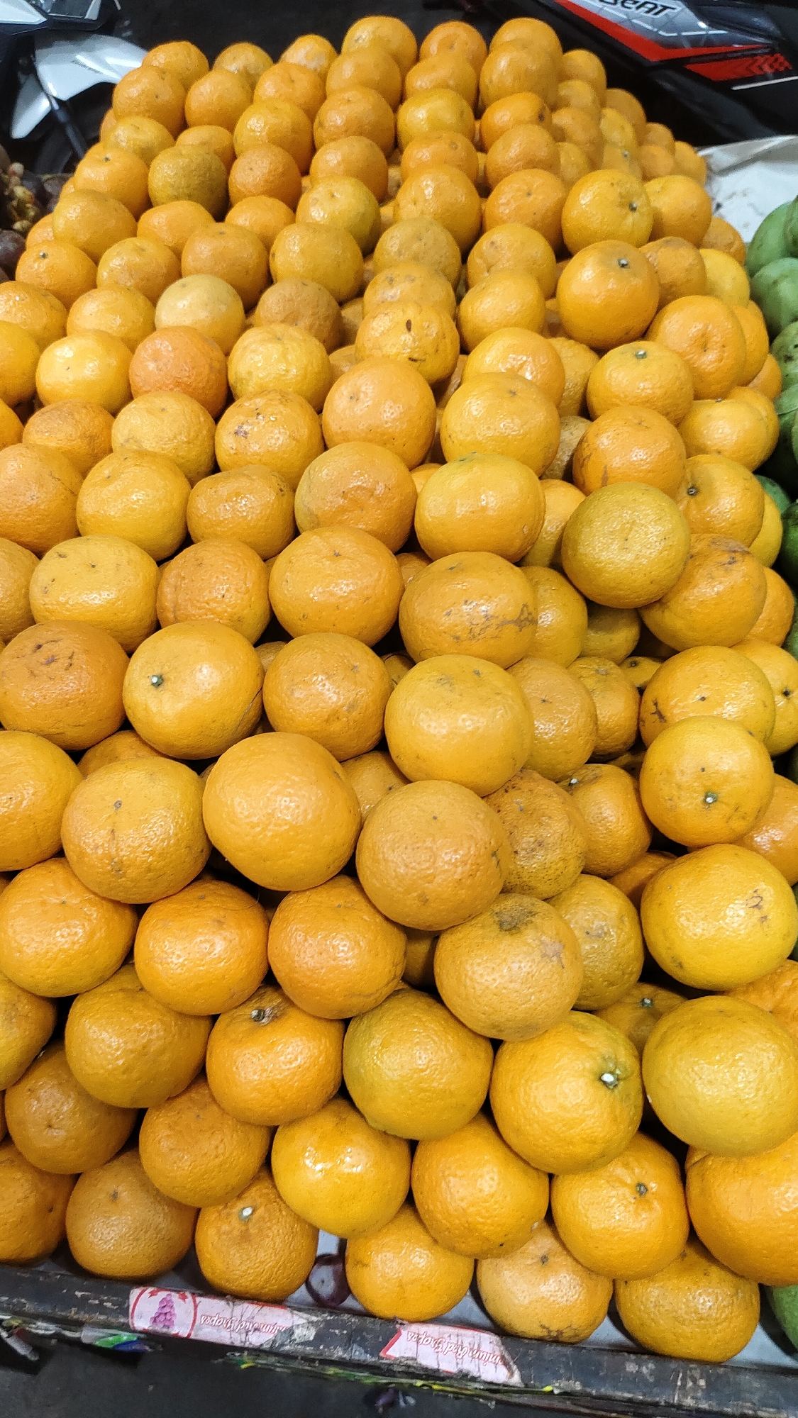 A close-up, eye-level, full shot captures a large, tiered display of oranges at a market stall in Malang, Indonesia. The oranges are predominantly bright orange, with some exhibiting slight variations in hue and texture, indicating they are fresh produce. They are piled high in a pyramid-like fashion, creating a visually dense composition. The lighting appears to be ambient, likely from the market's overhead illumination, casting subtle shadows and highlighting the rounded forms of the fruit. The background is blurred, but hints of other produce and a dark, possibly wooden, counter suggest a bustling marketplace environment. The image focuses solely on the oranges, with no people or specific actions visible. The overall impression is one of abundance and freshness.