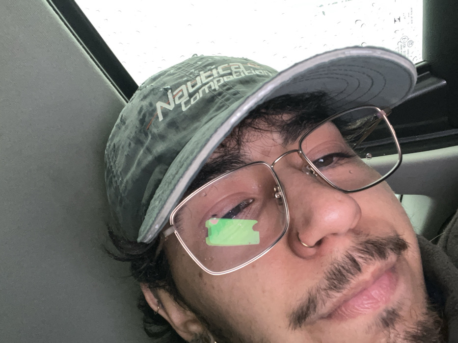 The image displays a tilted, close-up photograph of an individual inside a vehicle in Ushuaia, Argentina, during rainy weather. The subject, exhibiting masculine features, dark hair, a mustache, and beard stubble, wears a dark green baseball cap with a "Nautica Competition" logo, gold-rimmed rectangular eyeglasses, a gold hoop nose ring, and a silver earring in the left ear. Water droplets are visible on the cap and eyeglass lenses. A prominent bright green, irregularly rectangular reflection is present on the subject's right eyeglass lens. The vehicle's interior ceiling or trim is dark gray. Through a vehicle window on the right side of the frame, numerous water droplets adhere to the glass, confirming active precipitation against an overcast sky.