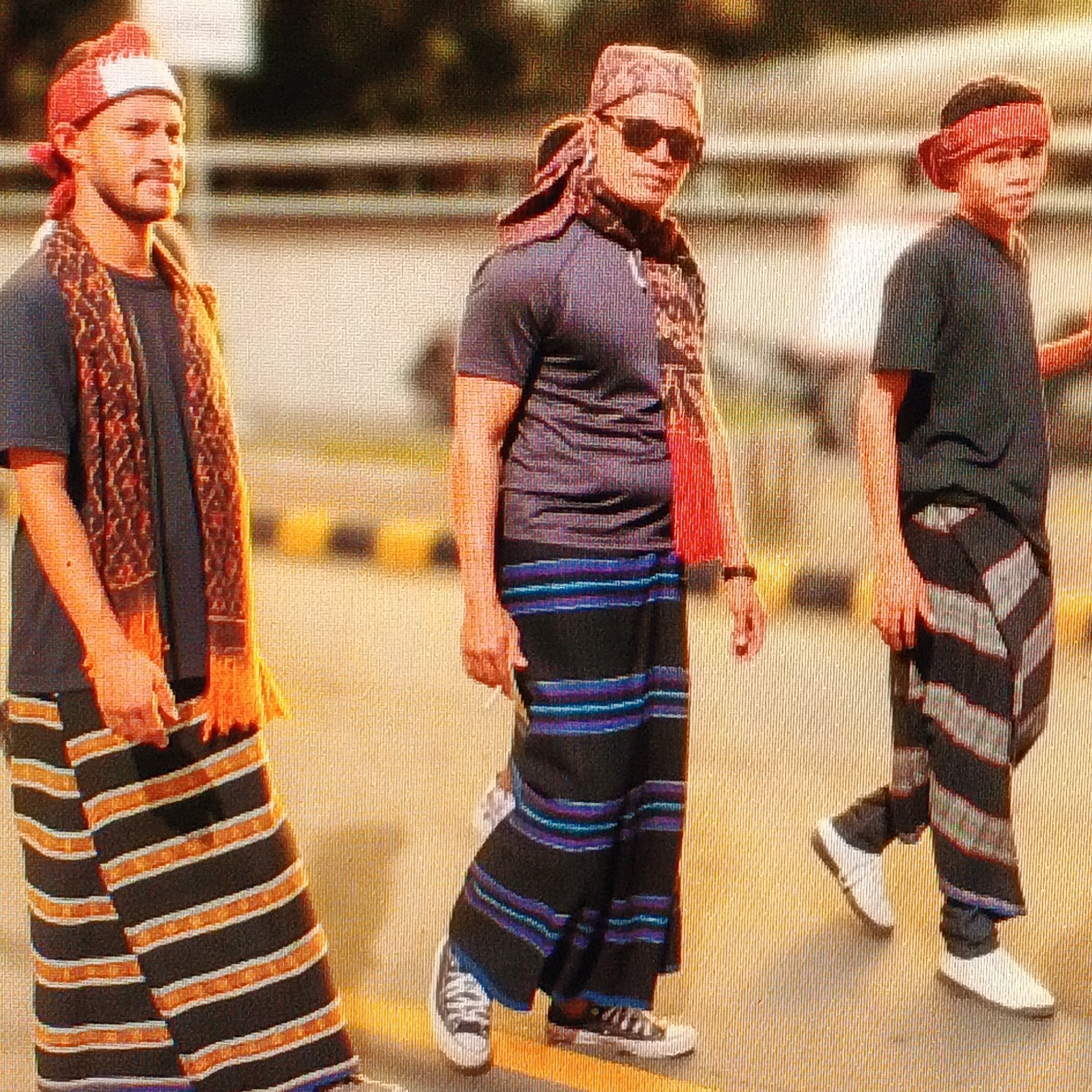 The image depicts three individuals walking outdoors, likely in the city of Ende, Indonesia. The first individual, on the left, is a man wearing a red and white head covering, a patterned scarf draped around his neck and shoulders, a dark t-shirt, and a sarong with horizontal stripes of black and orange. He is looking to the right. The second individual, in the center, is also a man wearing a patterned head covering, sunglasses, a dark t-shirt, and a sarong with horizontal stripes of black, blue, and purple. He is looking down and to the right. The third individual, on the right, is a younger person, possibly a boy, wearing a red head covering, a dark t-shirt, and a sarong with diagonal black and white stripes. They are looking to the left. All three individuals appear to be walking in the same direction. The environment suggests an urban setting with a paved surface and a blurred background that hints at buildings or other urban structures. The lighting indicates it is daytime, with a warm, golden hue over the scene, possibly due to the sun's angle or post-processing.
