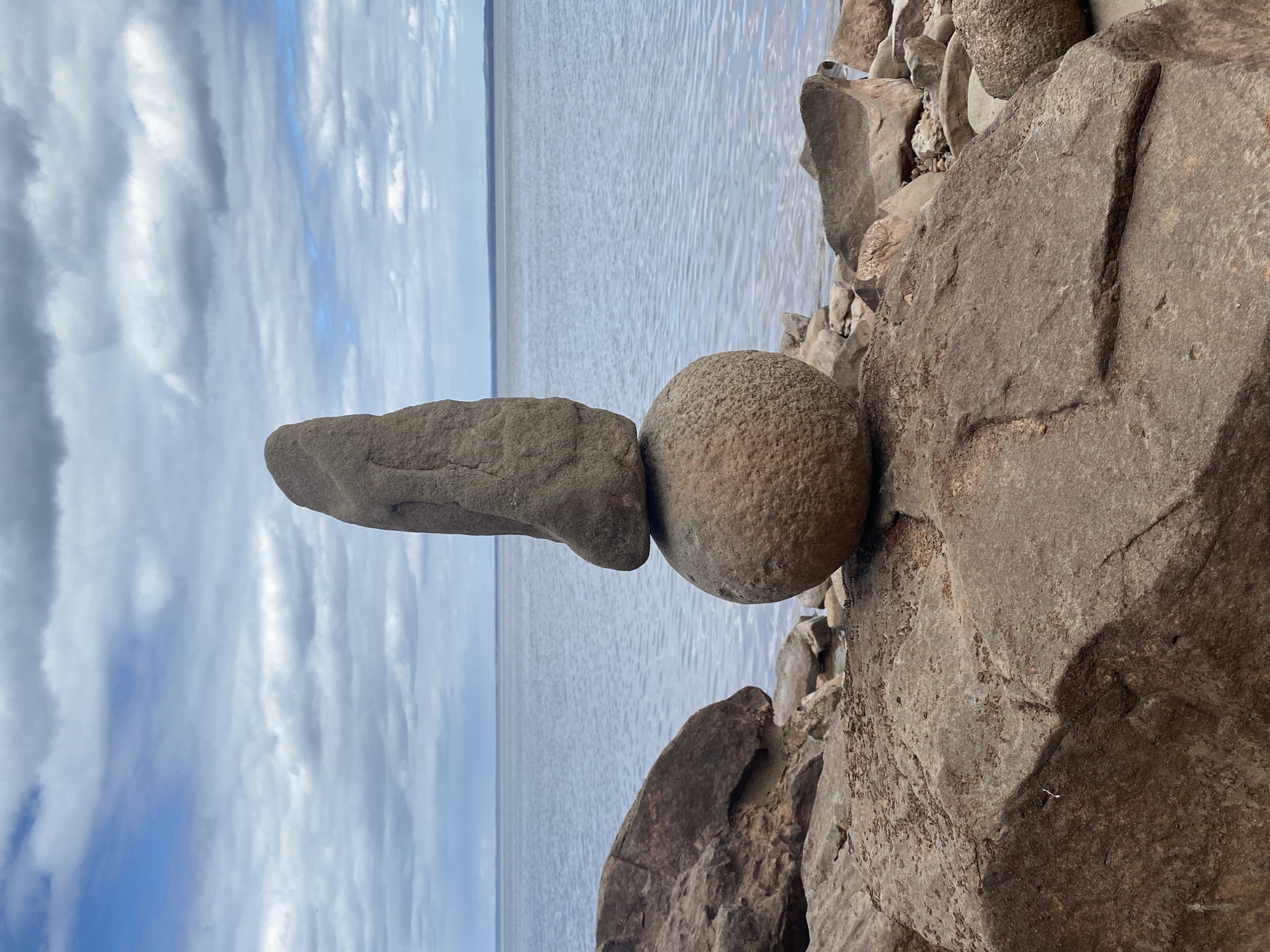 A stone sculpture, with a horizontally oriented, elongated rock balanced atop a round stone, sits in the foreground against a backdrop of a body of water and a cloudy sky. The round stone rests on a larger, rocky formation on the right side of the frame. The sculpture is positioned near the water’s edge. The location appears to be Noviy, Russia.