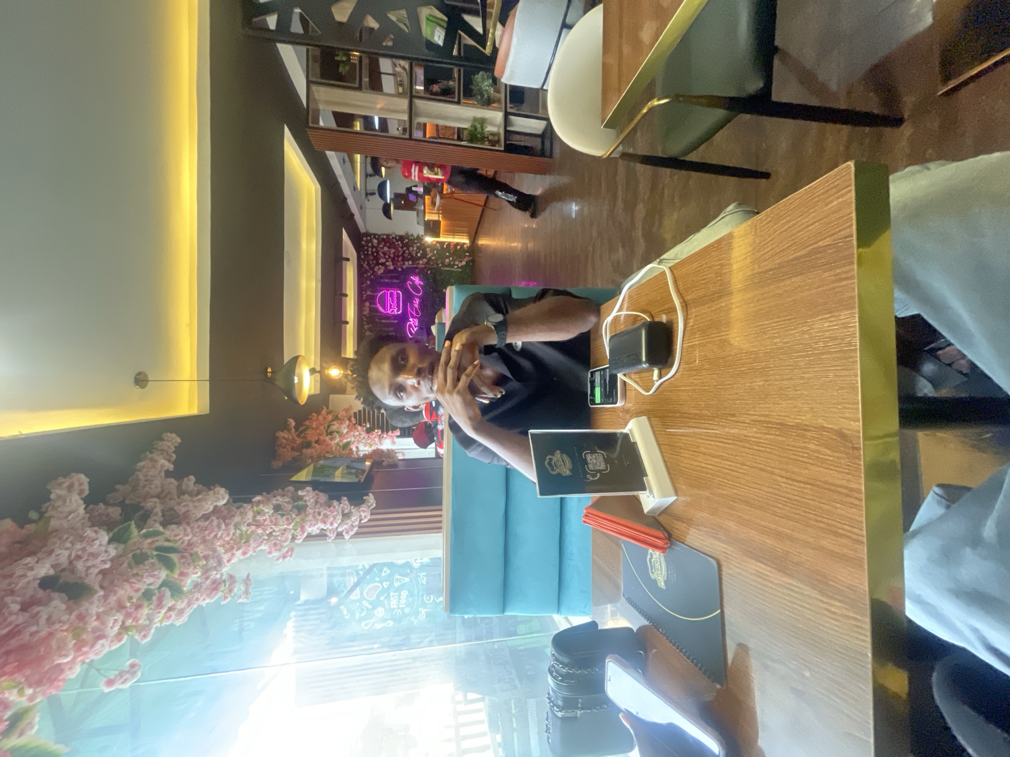 A man sits at a wooden table in what appears to be a restaurant or cafe in Adankolo, Nigeria. He is wearing a black shirt and is looking at the camera with his hand near his face. On the table are a menu, a phone, a wallet, and a charging block. There are flowers hanging from the ceiling. There is a neon sign that says "Rob Eze Cafe" in the background. A person is walking in the background as well.