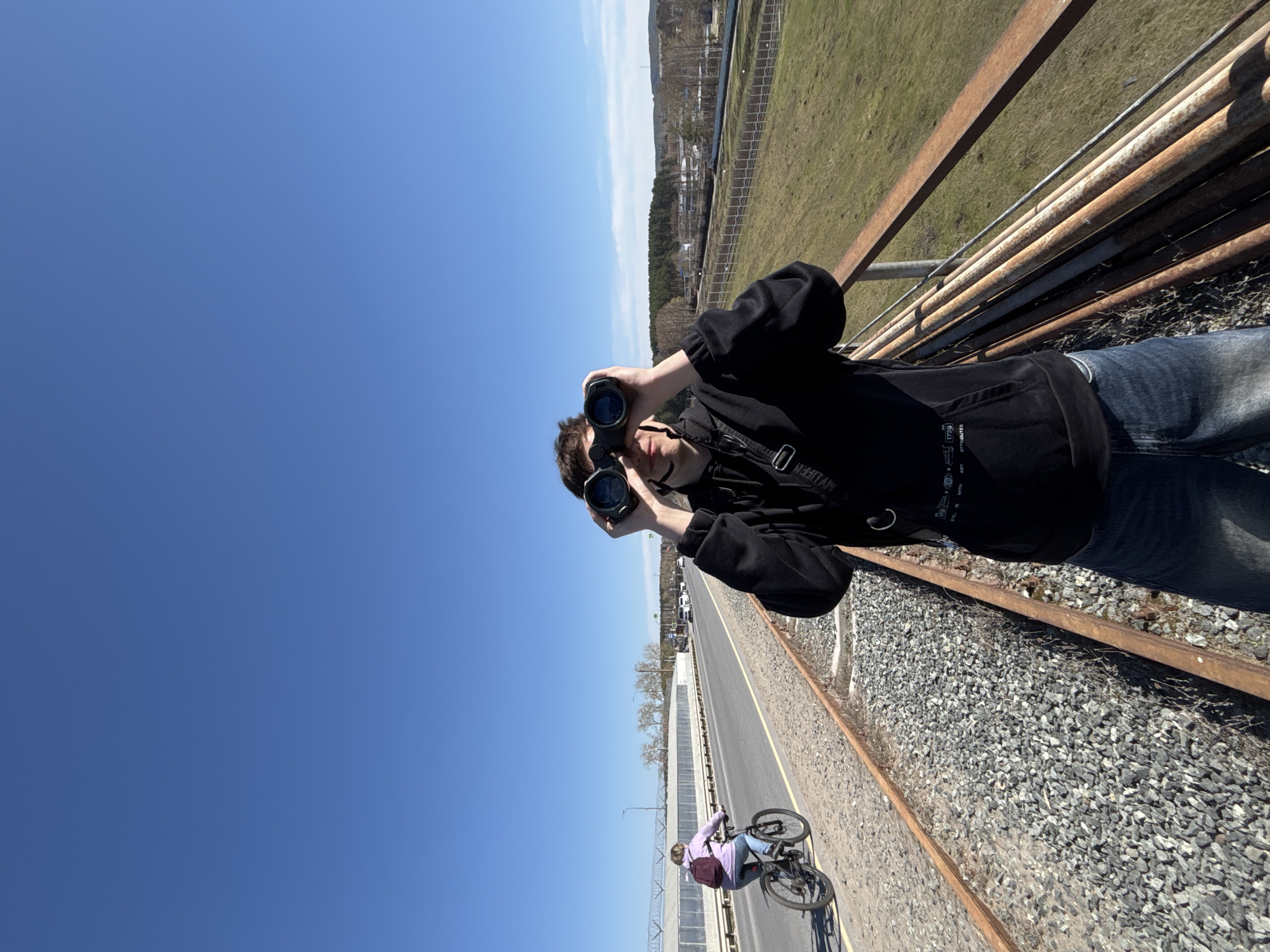 A person stands on a bridge over a road in Chaikovsky, Russia, holding binoculars. The road runs parallel to a railway track covered in gravel. A cyclist is seen on the road below. Beyond the road is a fence, trees, and distant buildings. The sky is clear and blue. The person is wearing a black hoodie and jeans. The bridge has rusty metal railings.