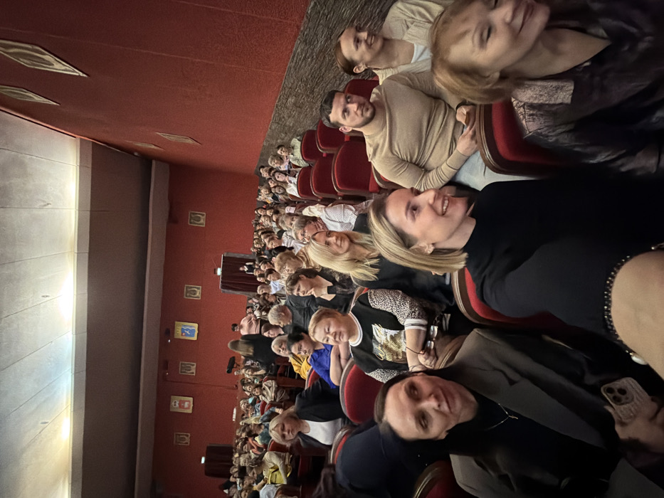The image displays the interior of a crowded auditorium or theater located in Chaikovsky, Russia. Rows of red upholstered seats are largely filled with an audience, predominantly adult women, with a few men interspersed. The attendees are seated, facing forward towards an unseen stage or presentation area, and are dressed in a mix of casual and semi-formal attire.
The room features dark red, textured walls, with a lighter, possibly fabric-covered, ceiling showing recessed lighting. Framed artworks or decorative plaques are mounted on the walls; some are rectangular with abstract designs, others are square with circular patterns. In the immediate foreground, from left to right: a woman with dark hair, wearing a dark blazer over a dark top, holds a light-colored smartphone with a textured case in her left hand, looking slightly to her left. Next to her, a woman with short blonde hair, wearing a black high-neck top, smiles broadly and looks towards her right. Behind them, a woman in a black and white patterned top looks forward with a slight smile. Further back and to the right, a man with dark hair and a beard, dressed in a light brown crew-neck sweater, looks forward, holding a small object in his right hand. Next to him, a woman with light brown hair in a light-colored jacket smiles towards her right. On the far right of the foreground, a woman with reddish-brown hair in a dark, shimmering top smiles to her right.
In the middle distance, several individuals are standing, including one woman with long dark hair seen from behind, near what appears to be a darkened entrance or a side section of the stage. The overall lighting in the room is moderate, suggesting an ongoing event or a moment before a performance.