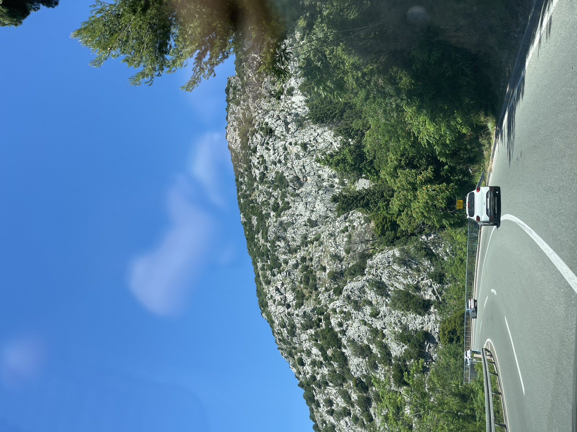 A wide, curving asphalt road, with white lane markings, passes through a mountainous, green landscape under a bright blue sky. The road is bordered by a metal guardrail on the left and dense green trees and vegetation on the right. Several cars are visible on the road, including a white SUV in the foreground on the right lane, a silver sedan and a white hatchback further ahead on the left lane, and a white sedan in the distance. The mountainside on the left is covered with gray rocks and scattered green vegetation. The sky is clear with a few wispy clouds. The overall scene suggests a scenic drive through a natural environment.