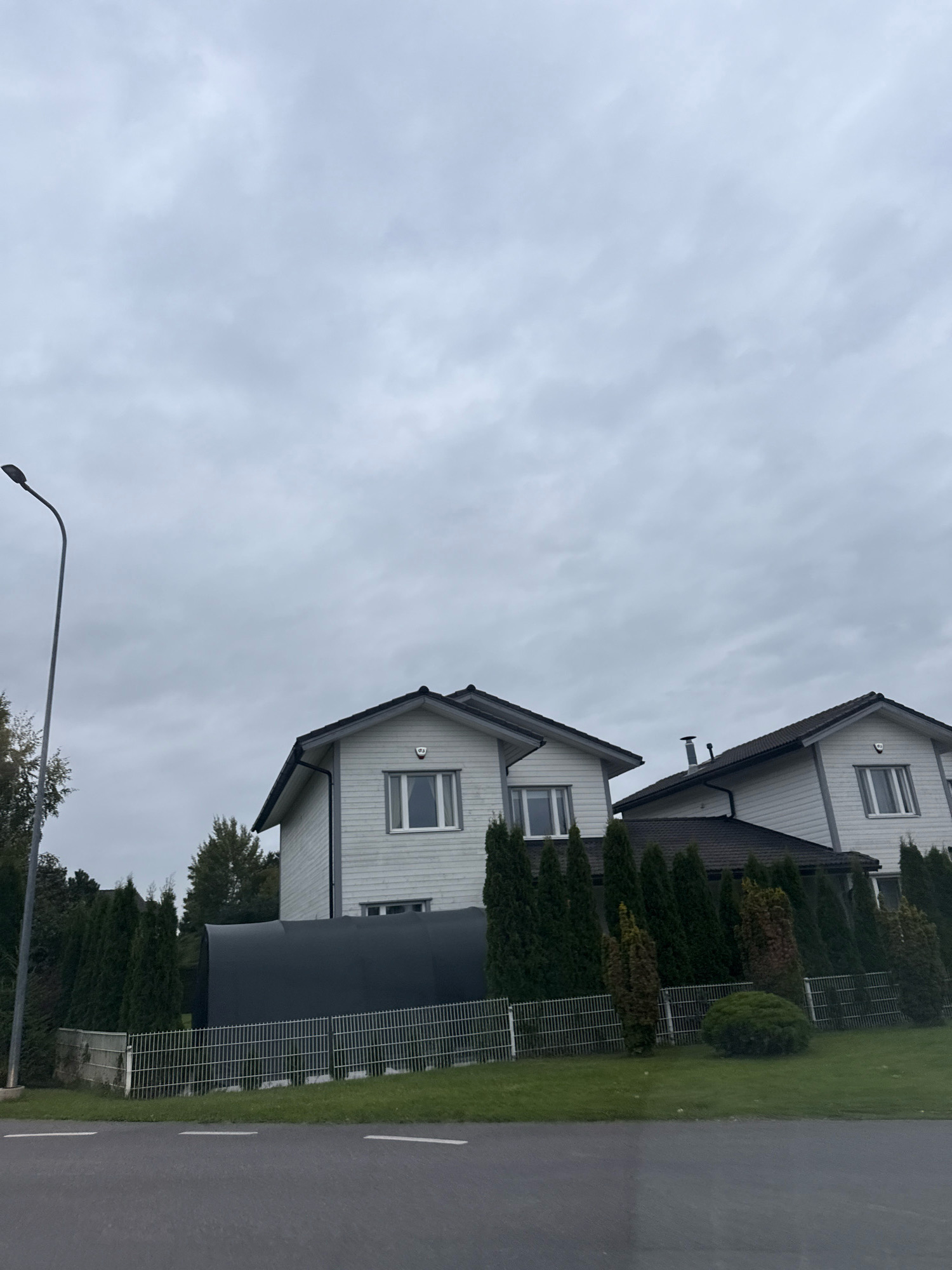 The image depicts a street view in Peetri alevik, Estonia, on an overcast day. In the foreground, a grey asphalt road with white lane markings is visible. Behind the road, a green lawn is bordered by a metal fence. Beyond the fence, a dark, curved structure, possibly a large storage unit or tank, is situated. Two modern, light-colored houses with dark roofs and white siding are visible in the background. The houses have multiple windows, and one of them has a security system logo above a window. A tall, grey street lamp with a curved arm stands to the left of the frame, near a cluster of evergreen trees and other foliage. The sky is completely covered in grey clouds, indicating a cloudy or possibly rainy day. There are no people or animals present in the scene.