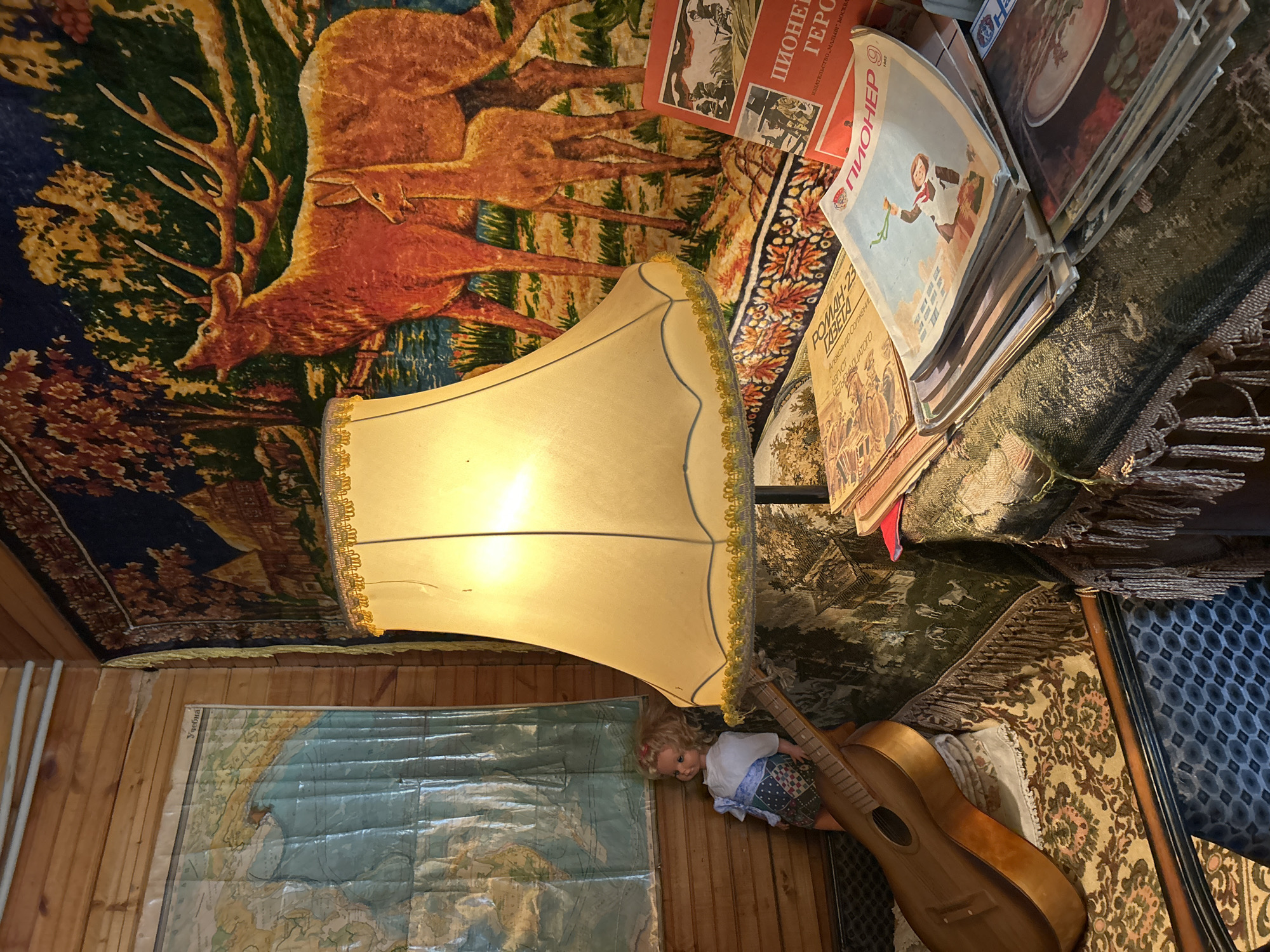 The image shows a cluttered interior, possibly a room in a house. The ceiling is covered with a tapestry featuring deer. A lamp with a beige lampshade dominates the center of the image. A stack of books and magazines rests to the side of the lamp, with titles like "ПИОНЕР ГЕРО" and "РОМАН-23 ГАЗЕТА". A world map is hung on a wooden wall. A doll and a guitar are present, resting against the wall. There is no discernible connection to the city of Moscow.