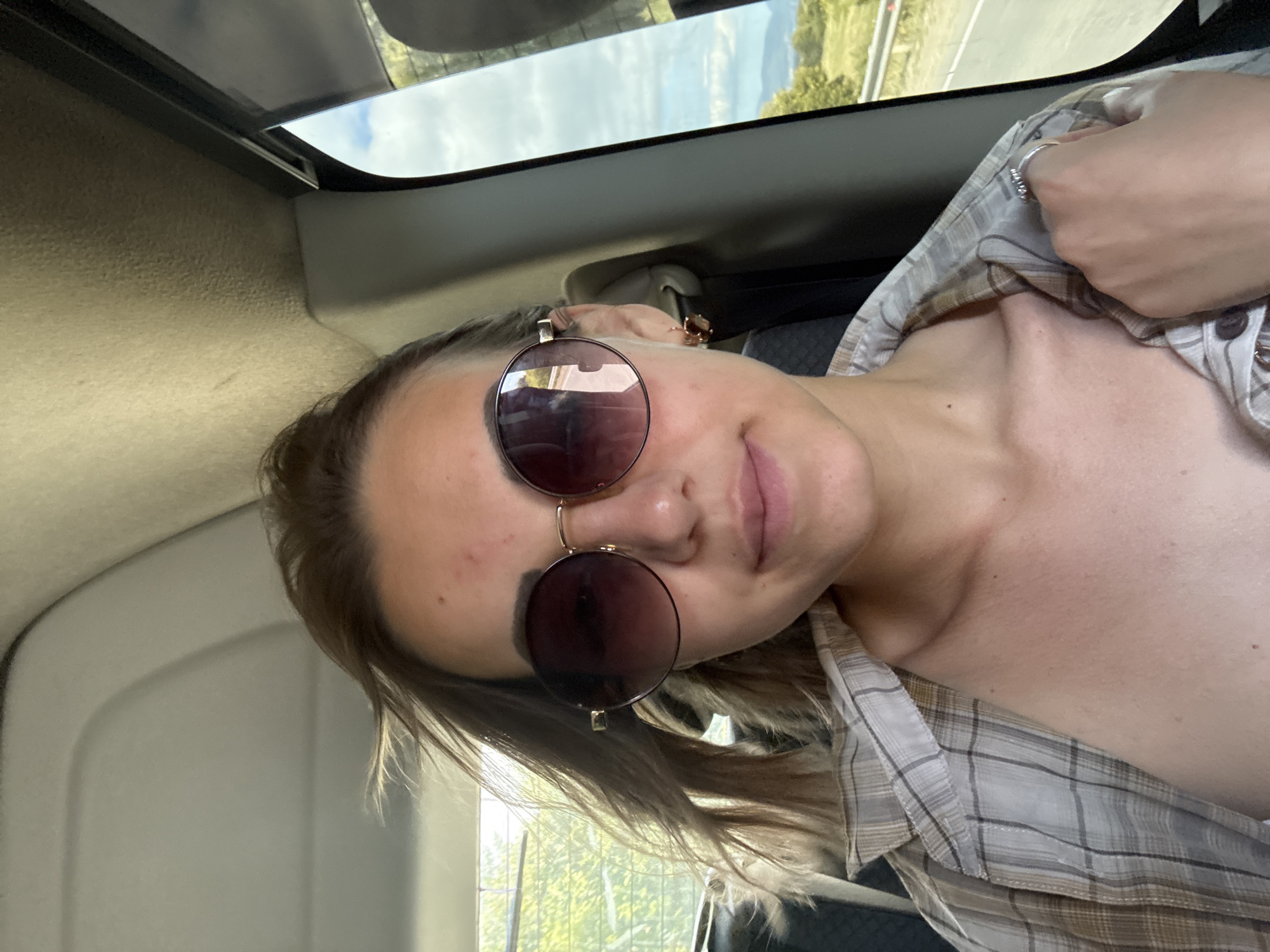 A young woman with shoulder-length brown hair and fair skin is shown in a close-up, selfie-style shot. She is wearing large, round sunglasses with gradient lenses and a tan and white plaid shirt, unbuttoned to reveal her upper chest and shoulders. Her hair is windswept, suggesting she might be in a moving vehicle. She has light makeup on, and a subtle smile plays on her lips. A small, gold earring with a dark charm is visible in her left ear. A silver ring is on the index finger of her right hand, which is resting on her chest. The background indicates she is likely inside a vehicle, with a view of trees, a road, and hills under a cloudy sky visible through the window. The lighting is natural and somewhat diffused, suggesting it is daytime.