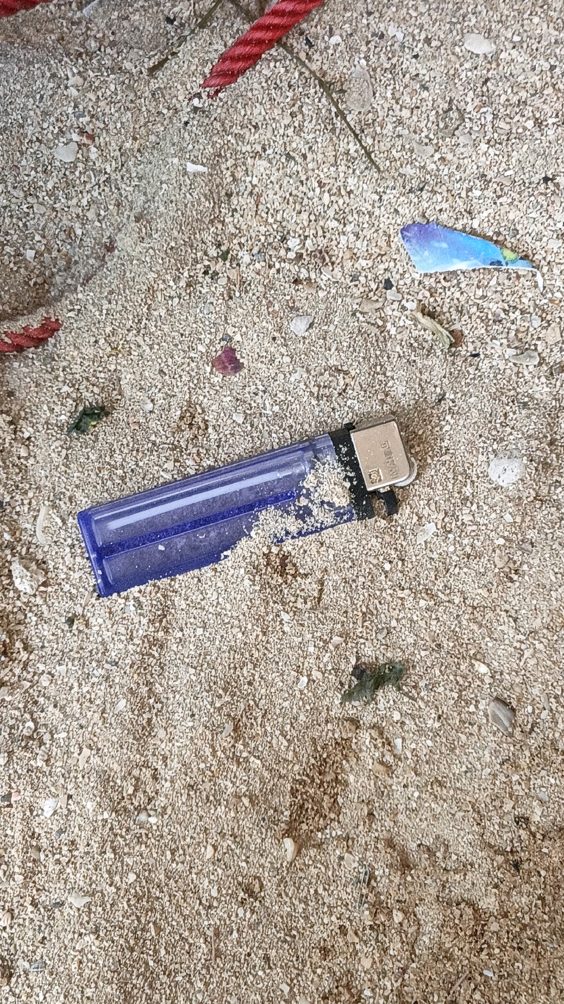The image depicts a close-up, overhead view of a sandy surface, possibly a beach. The sand is composed of fine grains and small shell fragments. Scattered across the sand are several items. A translucent purple plastic lighter, partially filled with sand, lies horizontally in the center of the frame. To the left and upper left, pieces of a braided red rope are visible, suggesting it is either partially buried or lying on top of the sand. In the upper right quadrant, a small, brightly colored piece of blue and yellow plastic debris is present. Several small pieces of green and brown organic matter, possibly seaweed or dried leaves, are also dispersed on the sand. The overall scene is one of beach detritus, with no discernible human presence or activity beyond the discarded items. The location is described as Cikal, Indonesia.