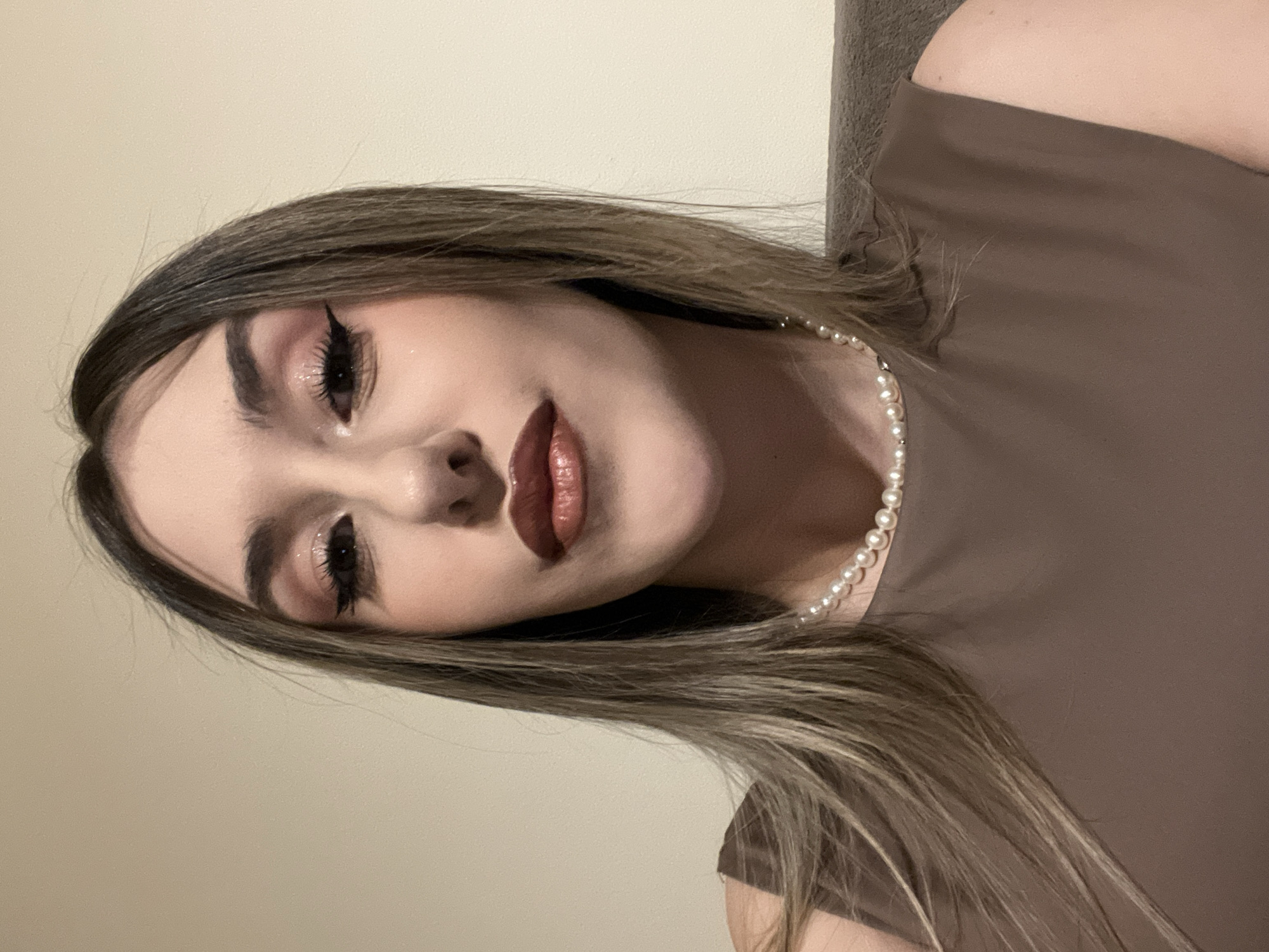 A woman with long, light brown hair poses for a portrait. She is wearing a brown top and a pearl necklace. Her makeup includes winged eyeliner, brown eyeshadow, and brown lipstick. The background appears to be a plain wall. There is no evidence to suggest that the image was taken in Sofia, Bulgaria.