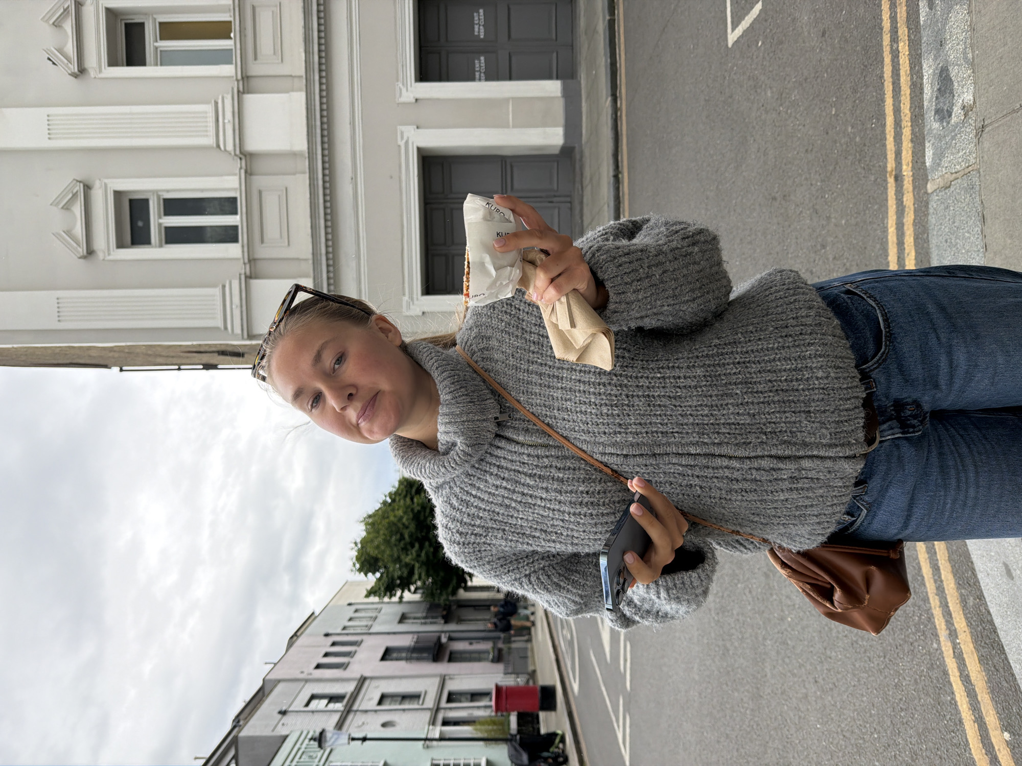 A young woman stands on a street in London, United Kingdom, holding a food item and a mobile phone. She wears a gray sweater, blue jeans, and a brown shoulder bag. Sunglasses rest on her head. The street has a double yellow line. Buildings line the street, with facades showing typical London architecture. A red postbox is visible. The sky is overcast. The woman appears to be taking a self-portrait or being photographed.
