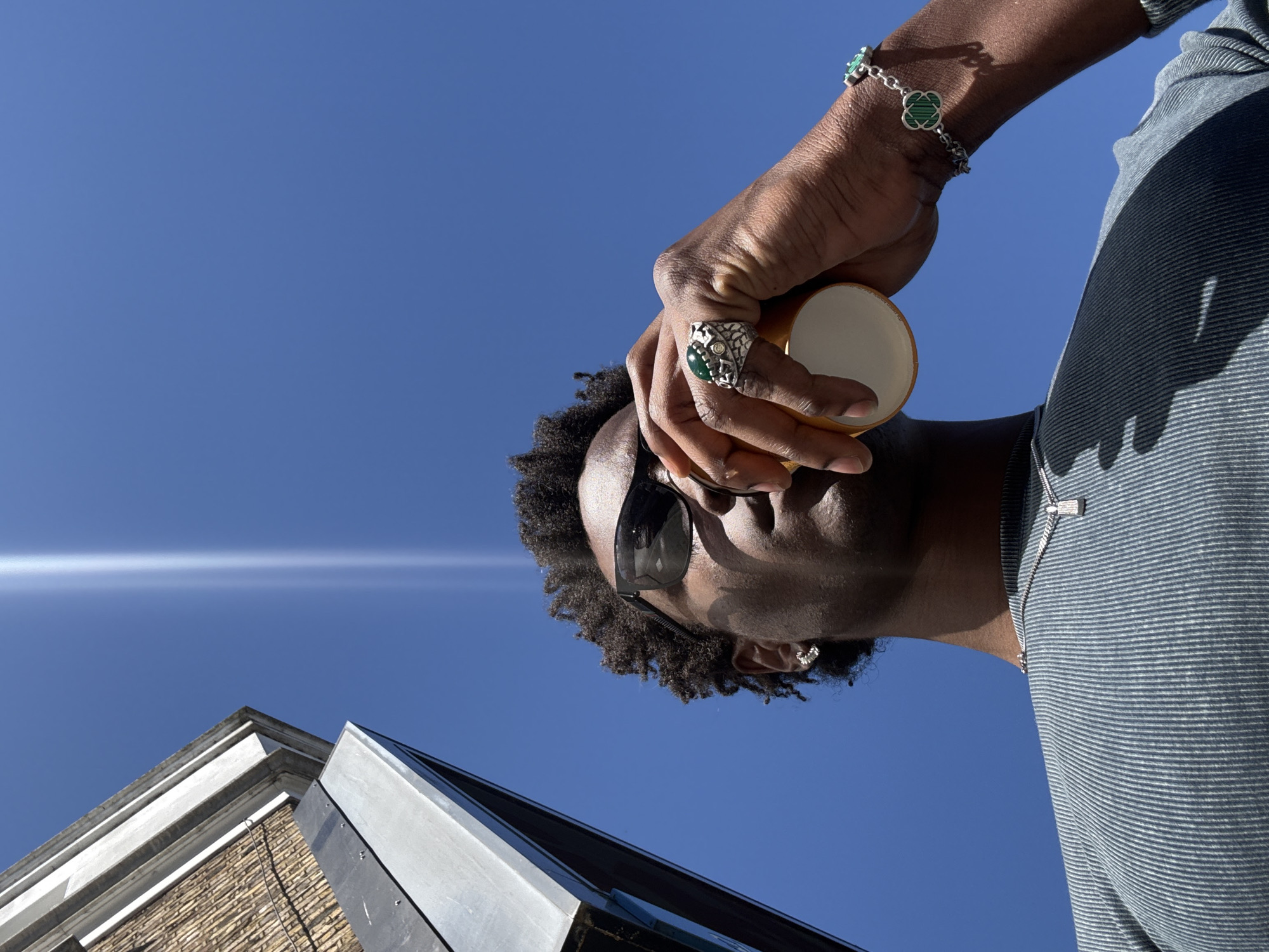 A low-angle shot depicts a dark-skinned person holding a cup against a bright blue sky, possibly in London, United Kingdom. The person is wearing sunglasses and has short, curly dark hair. They are adorned with jewelry, including a large ring with a green stone and a clover-shaped bracelet. The person is wearing a striped shirt. In the foreground, part of a building with brick and metal elements is visible. A bright, horizontal light flare appears in the sky. The perspective suggests the photo was taken looking upwards.