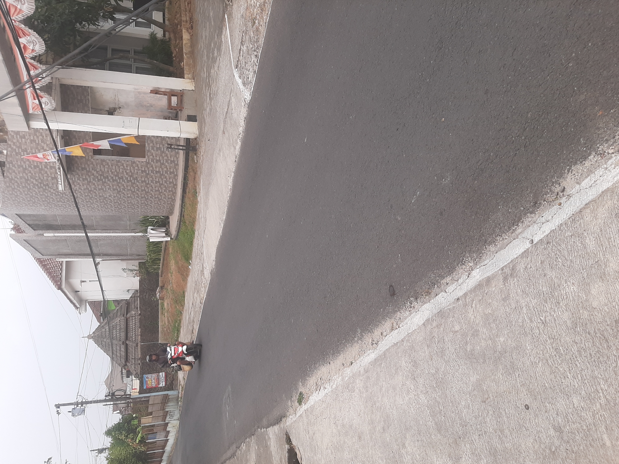 A person rides a white and red scooter on a paved road in Sumampir, Indonesia. The scooter is traveling away from the camera, and the rider is wearing a brown jacket and a helmet. The road is flanked by a concrete sidewalk on the left and a grassy area on the right. In the background, there are several buildings, including a house with a tiled roof and a modern building with a stone facade. A decorative roof structure, possibly part of a historical building or mosque, is visible behind a fence. Flags are displayed on the modern building. The sky is overcast.