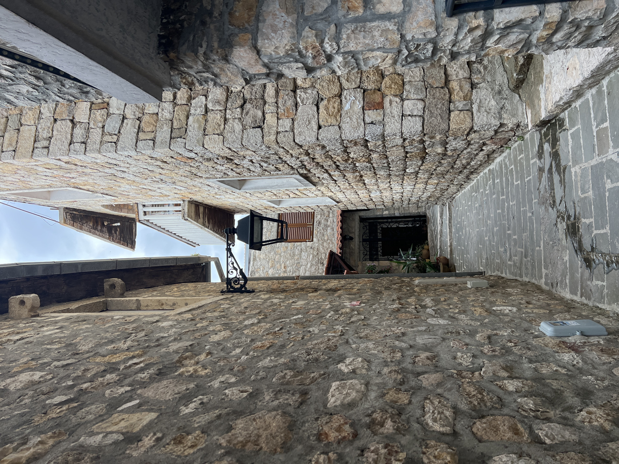 Captured from a low angle, the image depicts a narrow alleyway in Ulcinj - Ulqin, Montenegro. The path and the buildings' walls are constructed of stones. A decorative street light is present to the left. Vegetation is visible near the end of the alley. A utility box is present to the right. The sky is visible above.