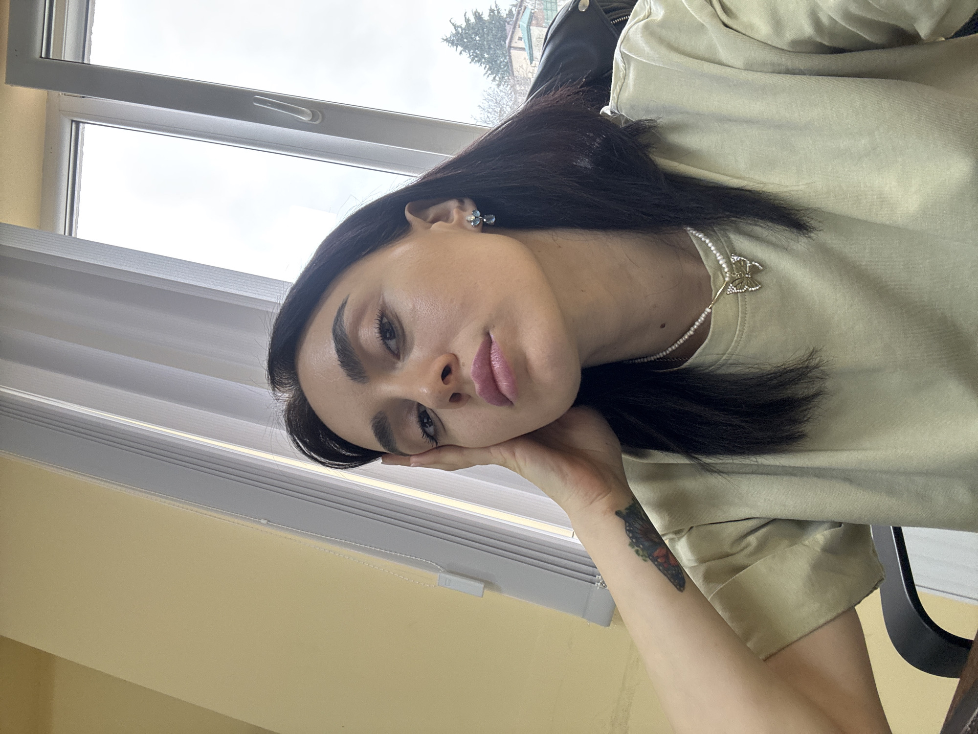 A close-up, eye-level portrait shows a single female subject indoors, facing the camera with a neutral expression. She has dark, shoulder-length hair and is wearing a light olive-green short-sleeved top, a white beaded necklace with a gold-colored butterfly pendant, and blue and gold drop earrings. A multi-colored butterfly tattoo is visible on her left forearm near the wrist. Her left hand is raised, with fingers resting against her left temple. She appears to be seated in a dark chair, partially visible behind her right shoulder. The indoor background consists of a light yellow wall on the left and a white-framed window. White vertical blinds are partially drawn over the left side of the window. Through the right side of the window, an overcast sky is visible above an exterior scene featuring bare tree branches, evergreen trees, and a building with a red-orange tiled roof, indicating an urban or suburban setting. The location is described as Tbilisi, Georgia.