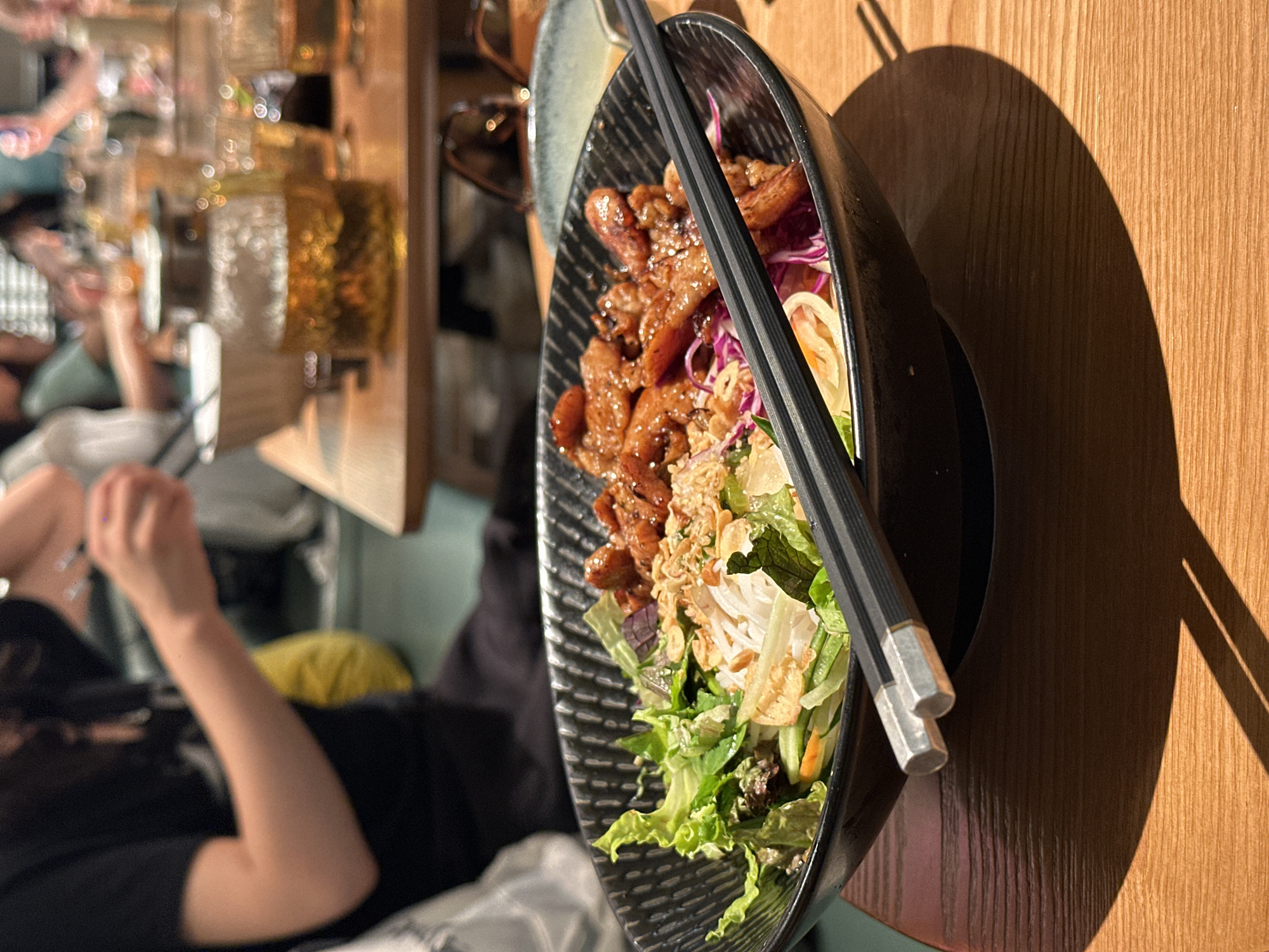 A close-up shot focuses on a black bowl filled with a Vietnamese dish, possibly Bun Thit Nuong, placed on a wooden table. The bowl contains rice noodles, grilled pork, fresh lettuce, shredded carrots, red cabbage, and fried shallots. A pair of black chopsticks with silver accents rests horizontally across the bowl. In the background, a glass teapot filled with amber liquid, several glasses, and other blurred figures are visible, suggesting a restaurant or dining setting. The lighting creates a warm ambiance with strong shadows cast on the table. The scene appears to be in a restaurant located in Futian District, China.