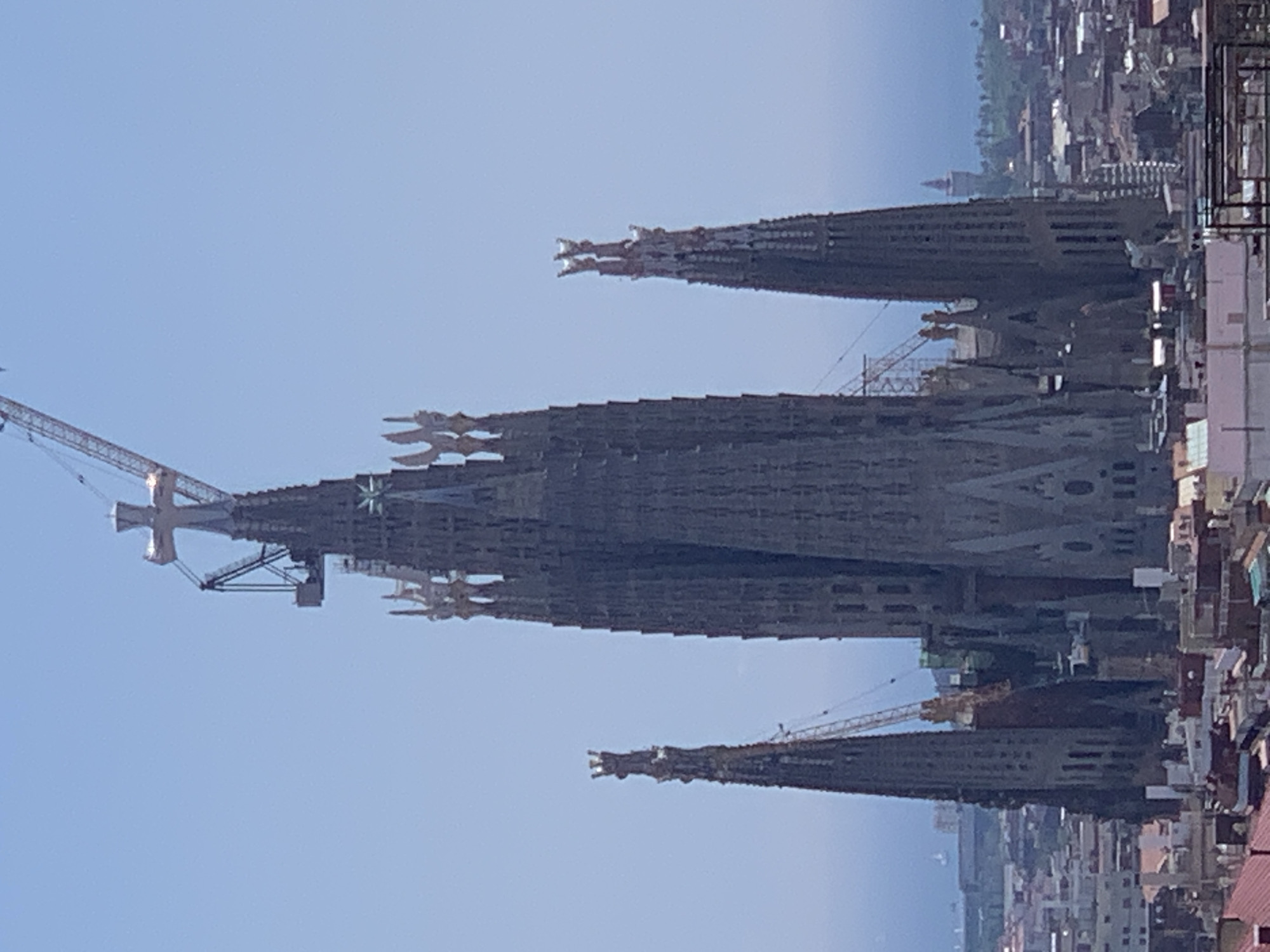 The image shows the Sagrada Familia basilica in Barcelona, Spain. Four prominent spires are visible, two with construction cranes attached. The basilica is surrounded by urban buildings. The sky is clear and light blue.