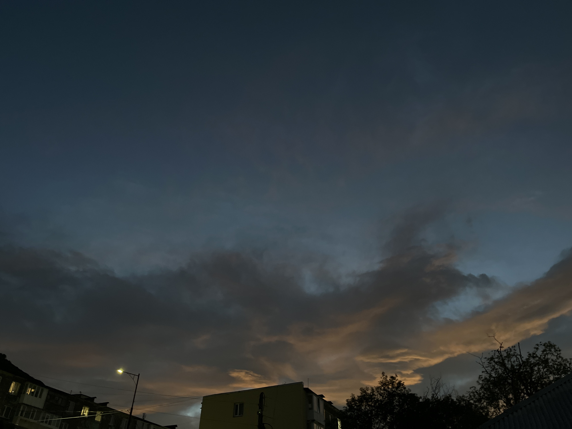 The image depicts a dusk or dawn sky over a city. The sky is a gradient of deep blue transitioning to lighter blues and greys, with streaks of warm orange and yellow light illuminating some of the clouds. In the foreground, silhouetted against the sky, are the rooftops of buildings and the dark foliage of trees. A single streetlamp casts a yellow glow, and lights are visible in the windows of the buildings, suggesting an urban environment. The scene is devoid of any visible people or specific actions. The overall impression is one of a serene, urban twilight. The location is identified as Petropavlovsk-Kamchatsky, Russia.