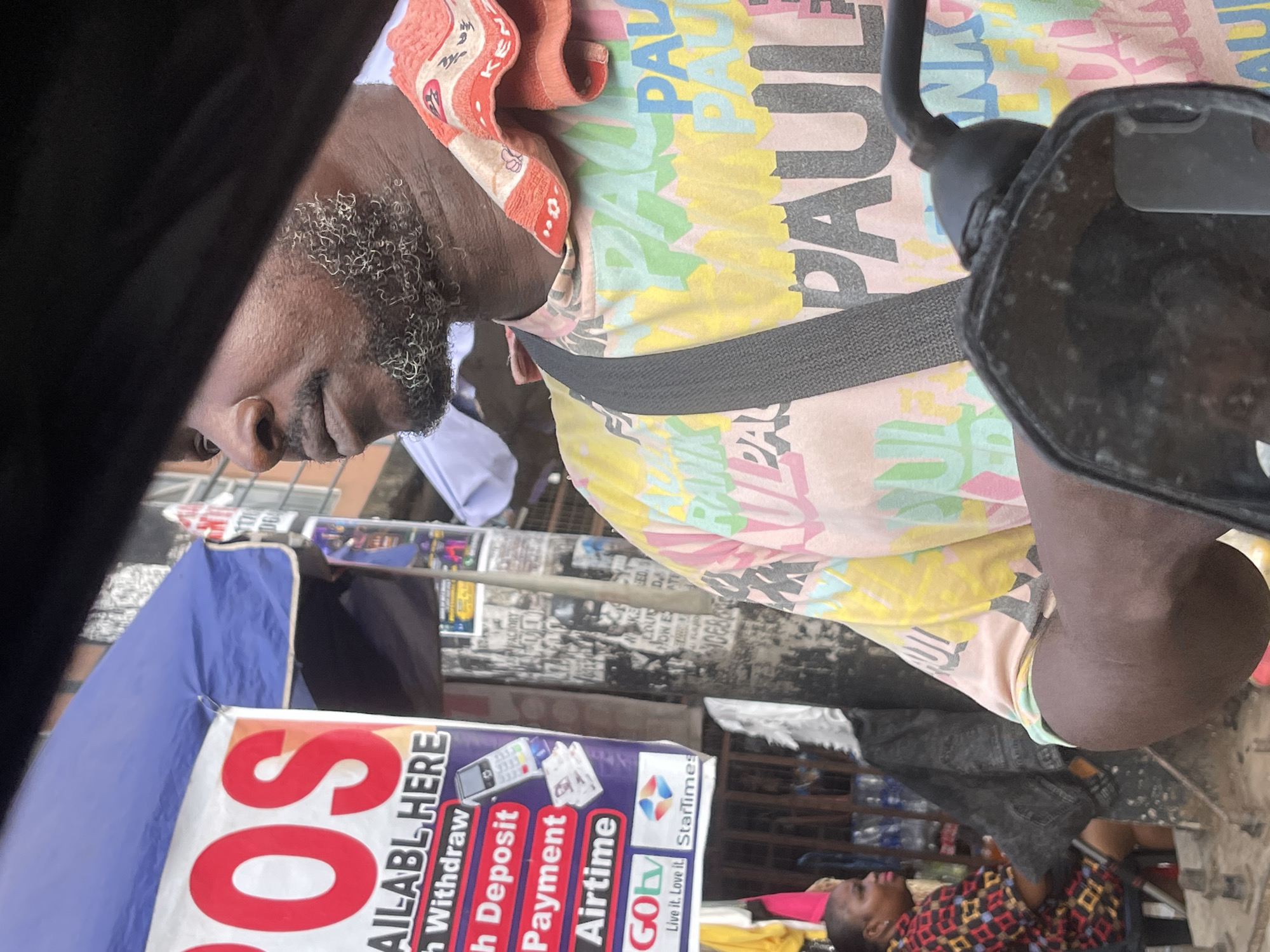 The image captures a bustling street scene in Onitsha, Nigeria, taken from the perspective of a vehicle, possibly a motorcycle, with a black handlebar or mirror visible on the right. The primary subject is a dark-skinned man positioned in the foreground, looking to his left. He has a beard with distinct white or grey curly hairs. He wears a vibrant, multi-colored shirt featuring the name "PAUL" repeatedly printed in various fonts and colors, and an orange patterned towel is draped around his neck.
In the mid-ground, a blue awning provides shade over a stall. Below it, a white banner is prominently displayed, advertising services with the partially visible red text "OS" and "AILABL HERE". Further text on the banner includes "Withdraw", "Deposit", "Payment", and "Airtime", suggesting mobile money or banking services. Logos for "GOtv" (with the tagline "Live it. Love it.") and "StarTimes" are also visible, indicating television services.
The background reveals a densely packed street environment typical of a market or commercial area. Various posters and flyers are plastered on walls, though most text is illegible, some visible words include "FACTORY", "LOUD DA", and "ATURJO". Another individual, possibly a child or small adult, wearing a patterned shirt, is seated or standing in the lower right background. The lighting suggests it is daytime. The image is rotated clockwise by 90 degrees.
