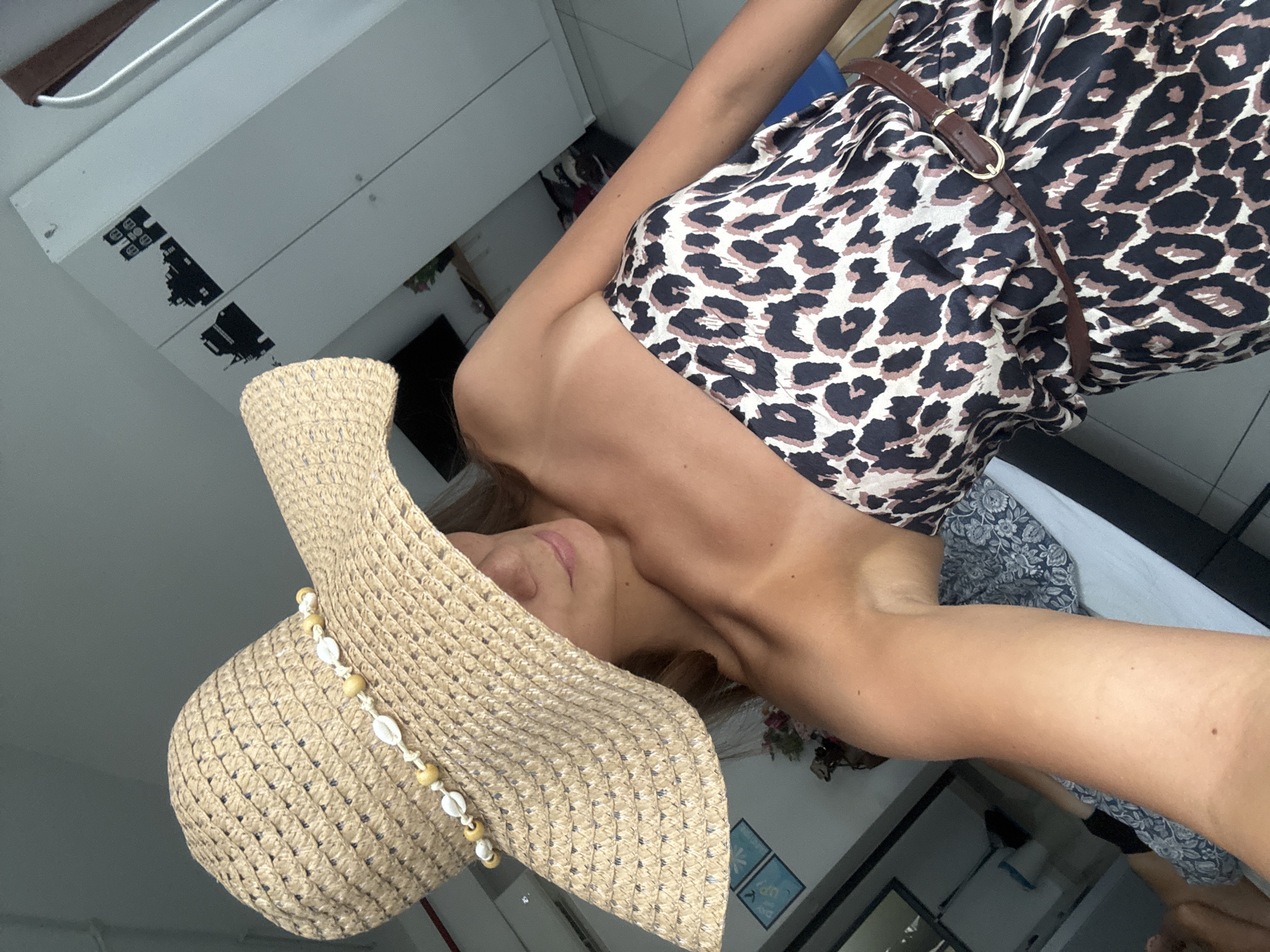 A woman wears a leopard-print dress with a brown belt and a large straw hat decorated with shells. She is positioned against a neutral-colored wall with a cabinet affixed. The photograph does not provide sufficient information to confidently identify the location as Kuala Lumpur, Malaysia.