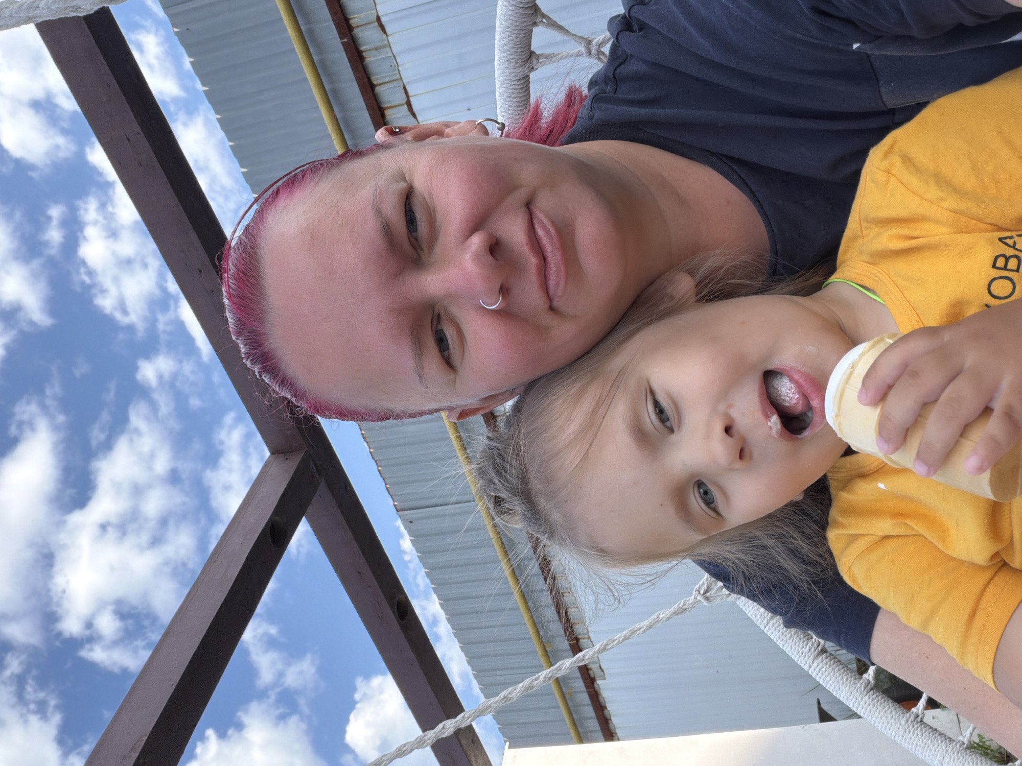 A woman with pink-dyed hair and a nose ring is holding a young child, who is eating an ice cream cone. They are sitting on a swing chair outdoors, with a wooden structure and a corrugated metal fence in the background. The sky is visible above with scattered clouds. The scene appears to be a casual, outdoor setting, likely in a residential area or park in the city of Chaikovsky, Russia.