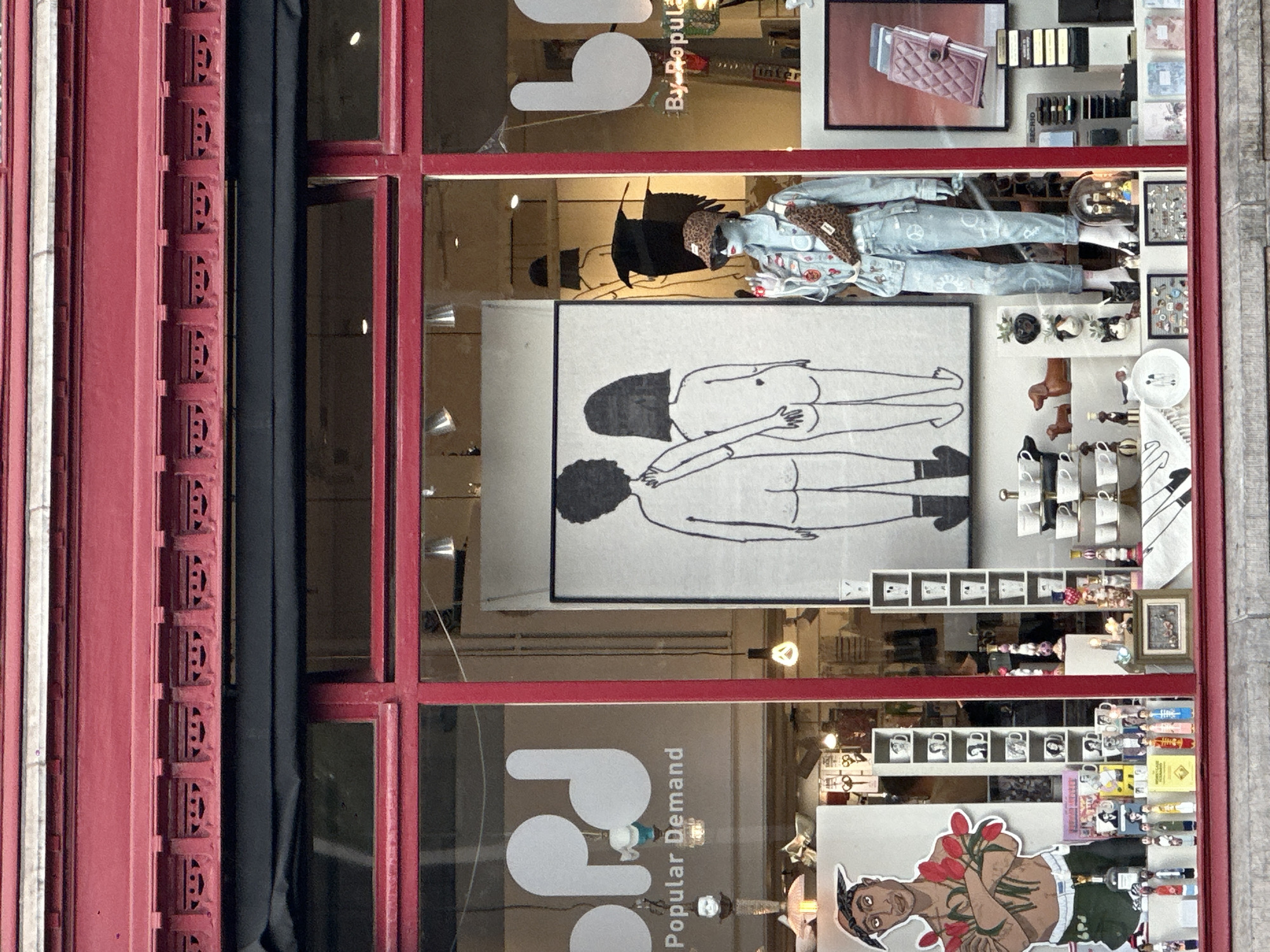 The image shows the storefront of a shop named "Popular Demand" in Amsterdam, Netherlands. The facade is painted a vibrant red with decorative molding above the windows. The shop window is divided into sections. On the left side, a large white logo with the letters "pd" is visible on a taupe background. Below the logo, "Popular Demand" is written in smaller text. To the right of the logo, a life-size cutout of a man with a bandana, holding red tulips, is displayed. The center section of the window features a large black and white drawing of two figures from behind, holding hands. A mannequin is dressed in a denim jacket and jeans, adorned with a leopard-print fanny pack and sunglasses. The mannequin is standing in front of a display of various items, including mugs, dog figurines, and small decorative objects. The right side of the window has another "pd" logo, and further to the right, a framed picture of a quilted pink item is displayed, along with stacks of books and notebooks. The window also contains hanging lamps and other assorted merchandise.
