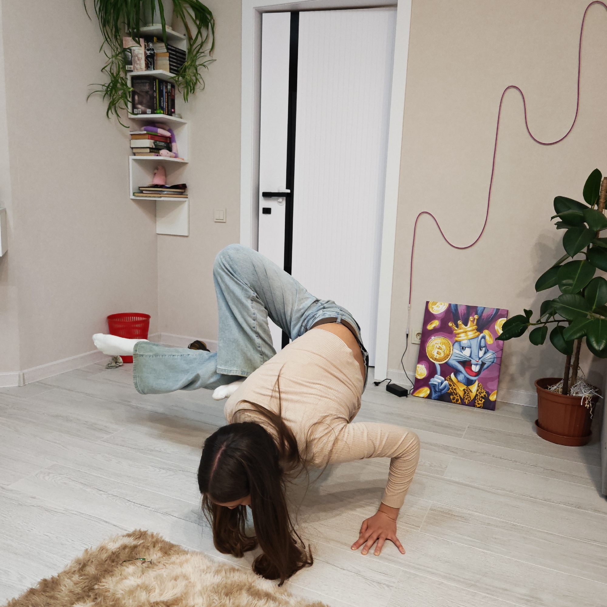 A young female with long brown hair is performing a gymnastic pose in an indoor environment, likely a residence. She is bent backwards with her hands and feet on the floor. She is wearing light blue jeans and a light beige long-sleeved shirt. A beige, shaggy rug is partially visible in the bottom left corner. The floor is light grey wood-look tile.
In the background, there is a white door with a black vertical accent and a black handle. To the left of the door is a corner shelf holding books and plants. A red trash can is visible in the background. To the right of the door, a cord runs vertically in a wavy pattern along the wall. A framed print featuring a cartoon rabbit with a Bitcoin theme leans against the wall near a potted plant.
The scene is likely located in Rostov, Russia, based on possible cultural or architectural features that are consistent with that location.