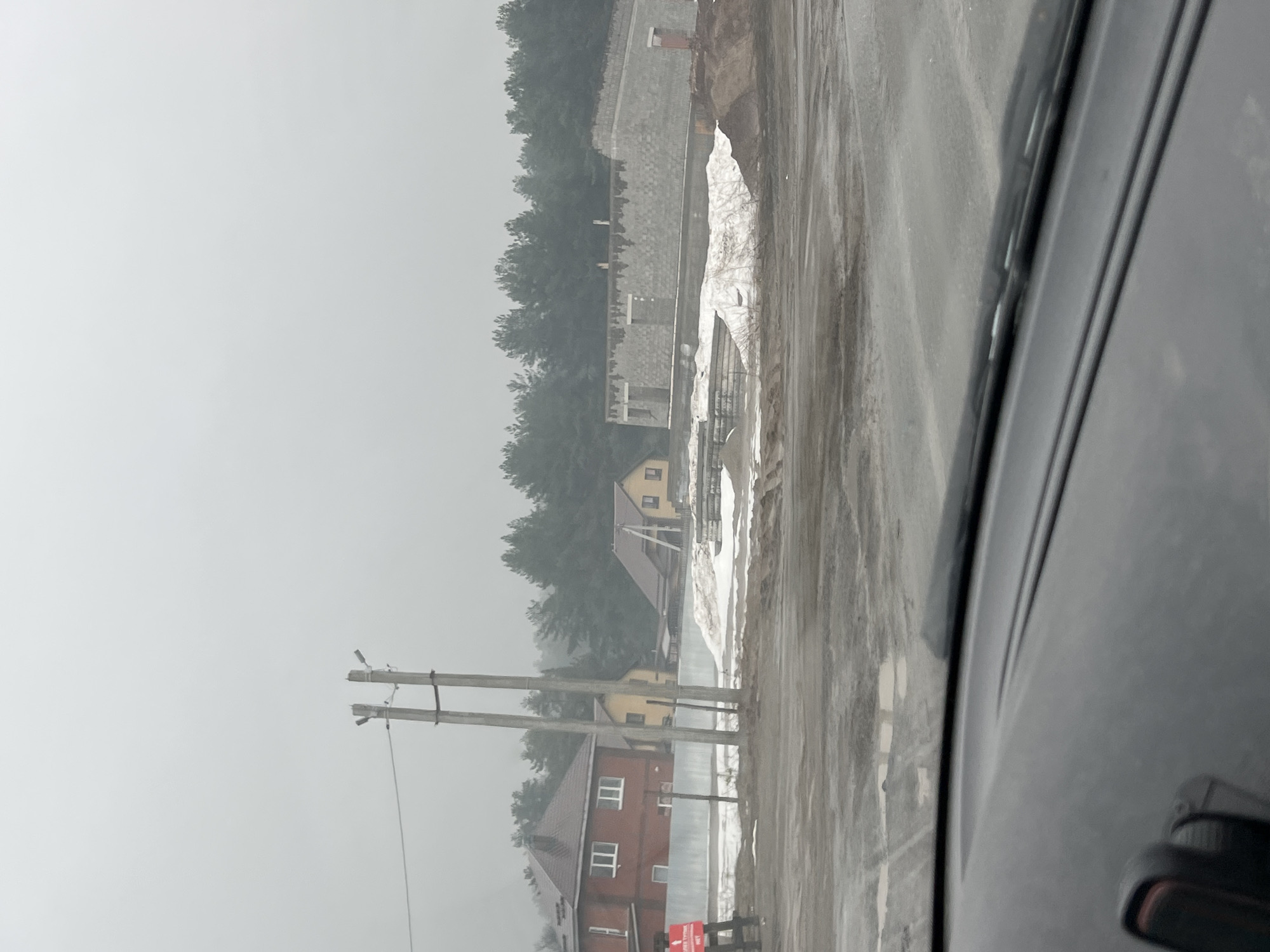 The image shows a view from inside a vehicle, likely a car, looking out onto a street scene in what appears to be the city of Chaikovsky, Russia.
**Environment:** The weather seems overcast and hazy, possibly snowy or foggy. The road appears wet and dirty with tire tracks visible. The vegetation indicates it might be a suburban or rural area on the outskirts of the city.
**Objects:** The most prominent objects include:
* Power lines on the left side of the frame.
* Several residential buildings, including what looks like two-story houses of different colors (red, yellow). They have typical sloped roofs and windows.
* A large wall or structure under construction behind the buildings. There's snow accumulation on top of it.
* The dashboard and part of the side of the vehicle the photo is taken from, including the side mirror.
**Subject:** There's a suggestion of human presence with the houses, but no people are clearly visible. The primary subject is the scenery of a Russian settlement.
**Location:** Given the architectural style of the houses, the infrastructure (power lines), and assuming the location is Chaikovsky, Russia, this scene likely depicts a residential area on the city's periphery.
**Actions/Interactions:** No specific actions or interactions are occurring in the image beyond the implied movement of the vehicle.