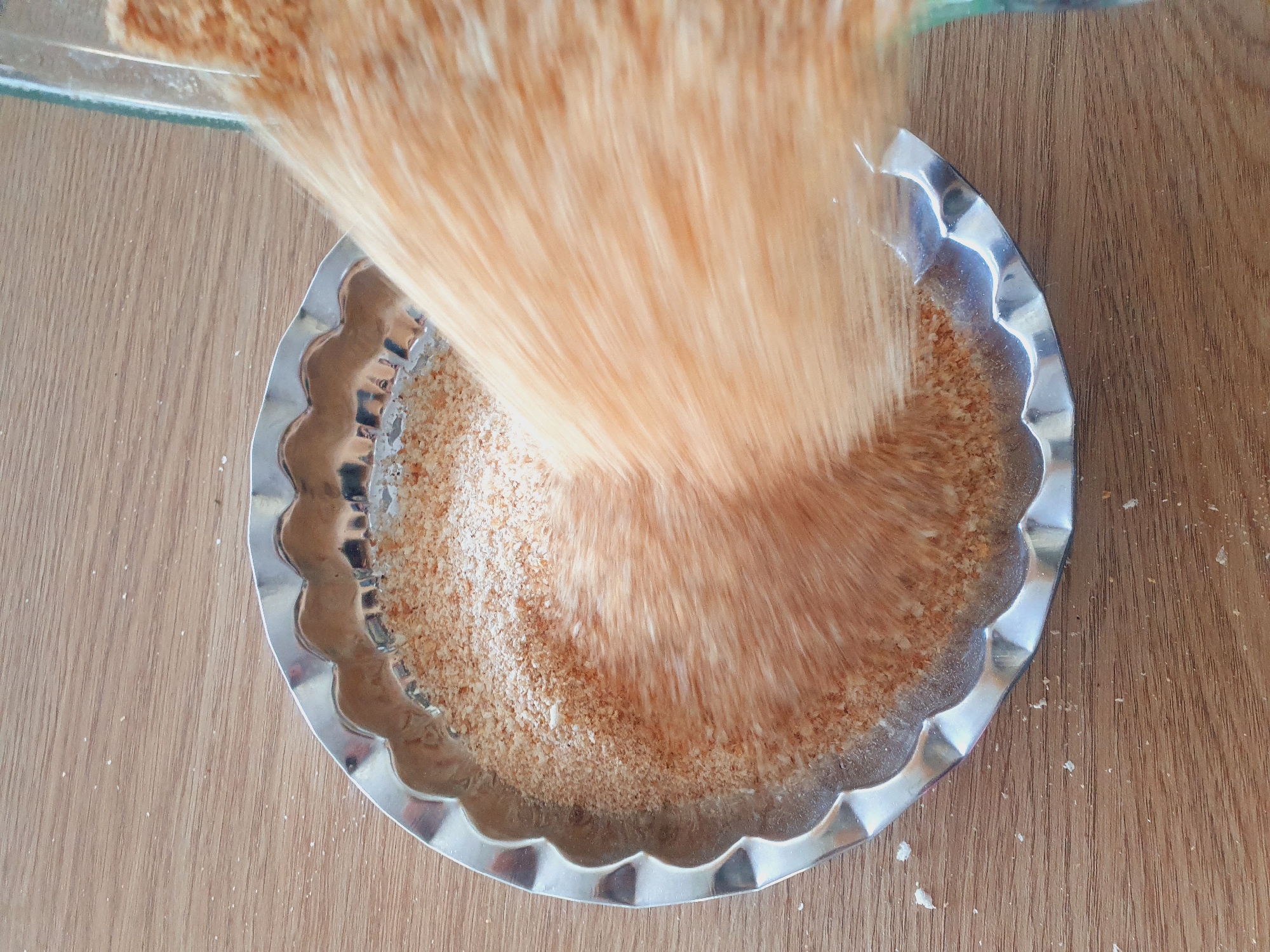 The image shows a close-up, top-down view of a fluted metal pie dish filled with breadcrumbs. A blurry stream of orange-colored breadcrumbs is being poured from a clear glass container into the dish, obscuring a portion of the dish and its contents. The dish is placed on a wooden surface, likely a table, with scattered white crumbs visible on the wood. The scene is set indoors, suggesting a kitchen environment, but no specific location in Kheyrabad, Iran, can be identified from the visual information provided. The lighting appears to be natural or overhead artificial light, casting subtle reflections on the metal dish.