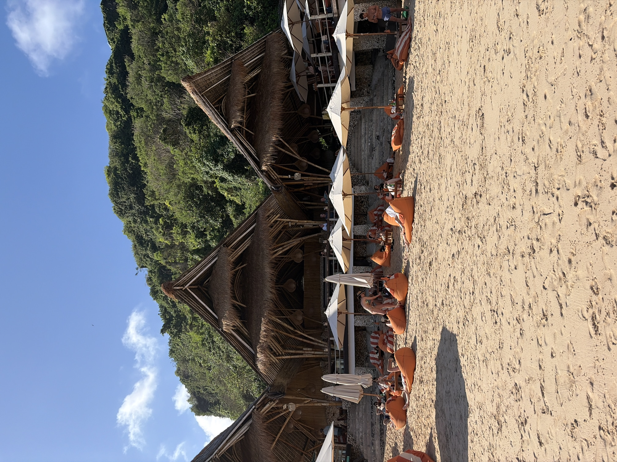 This image depicts a beachfront scene at a resort or beach club in Kuta Selatan, Indonesia. In the foreground, golden sand covers the beach with footprints visible. A collection of large, orange beanbag chairs and some striped loungers are scattered across the sand, many occupied by sunbathers. Several white beach umbrellas are open, providing shade. Behind the seating area, a tiered structure with bamboo accents and thatched roofs houses an open-air restaurant or bar. Steps lead up to this elevated structure. People are visible seated at tables and on the upper level of the building. In the background, a lush, green cliffside rises steeply, covered in dense vegetation. The sky is a clear blue with a few wispy white clouds. The overall impression is one of a relaxed, tropical beach destination.