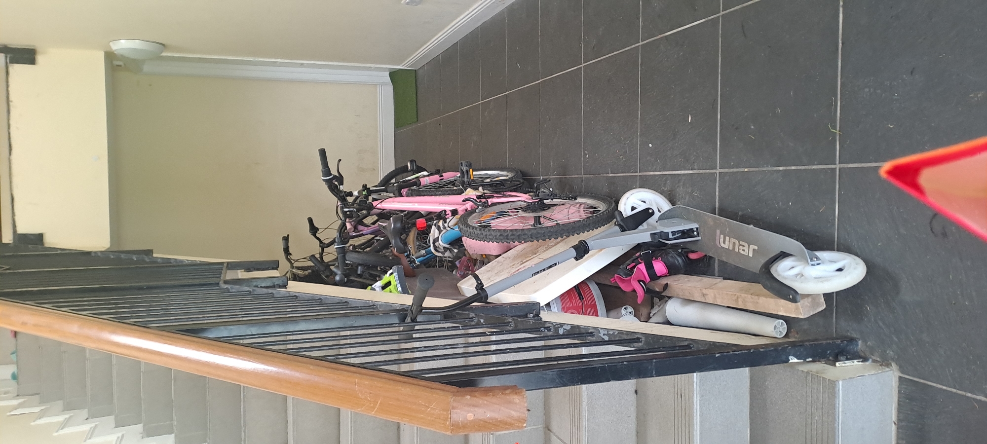 The image depicts a stairwell area in Castlebar, Ireland, where several bicycles and a scooter are stored. The scene is lit by ambient light, suggesting it is daytime.
In the foreground, a grey scooter with a black deck and white wheels is positioned diagonally across the tiled floor. The word "lunar" is visible on the deck. To the left of the scooter, a pair of pink and black rollerblades with buckles are placed on the floor. Behind the scooter and rollerblades, a collection of bicycles is leaning against the wall and railing. One prominent bicycle has a bright pink frame, black tires, and silver handlebars. Other bicycles are visible behind it, in various colors including black and possibly blue, with some showing green components.
The stairwell features grey concrete steps with a wooden handrail that runs alongside a black metal railing. The walls of the stairwell are painted a light, creamy yellow color. The floor is tiled with dark grey, square tiles. In the background, a white door frame is visible, and a white circular object is mounted on the wall. A small, green mat lies on the floor near the doorway. The overall impression is one of clutter and disarray, with the vehicles and accessories occupying a significant portion of the shared space. There are no people visible in the image.