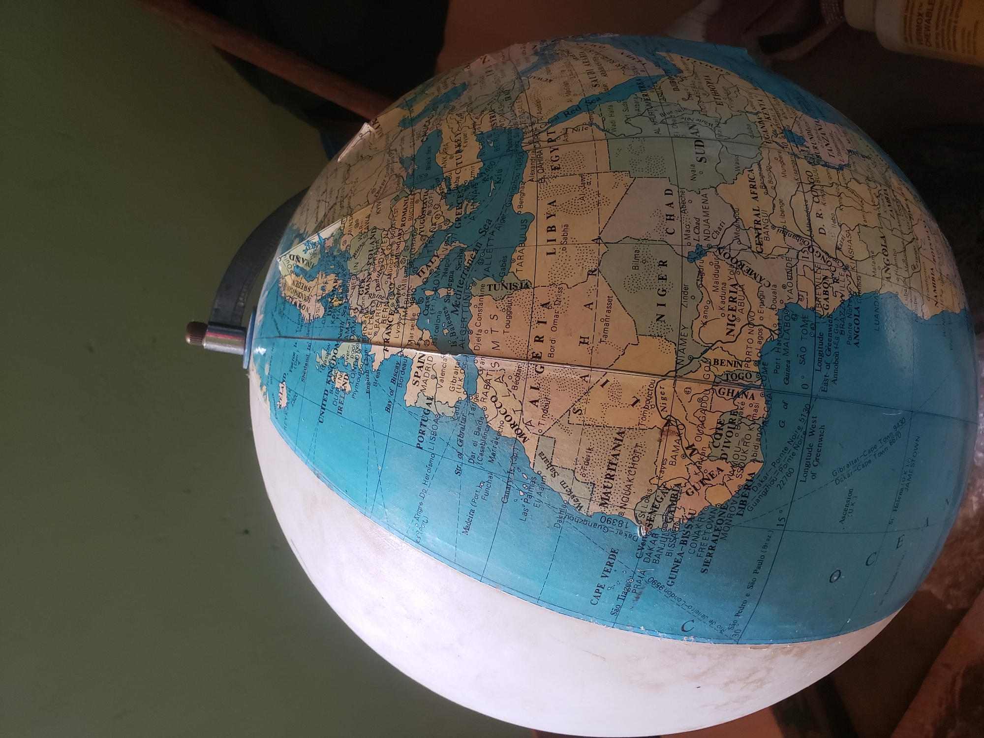 A close-up, slightly high-angle shot shows a portion of a vintage terrestrial globe mounted on a stand, set against a muted green, out-of-focus background. The globe itself is primarily blue and cream, with continents depicted in tan and cream colors, and country borders delineated by thin black lines. Text and place names are visible in dark lettering. Notable locations visible on the globe include parts of Europe (Ireland, United Kingdom, France, Germany, Spain, Portugal), Africa (Morocco, Algeria, Libya, Egypt, Mauritania, Senegal, Mali, Niger, Chad, Sudan, Ethiopia, Nigeria, Central African Republic, Cameroon, Equatorial Guinea, Gabon, Congo, Angola, Namibia, Botswana, South Africa, Zimbabwe, Mozambique, Tanzania, Kenya, Uganda), and the Atlantic Ocean.
Specifically, the city of Ajaokuta, Nigeria, is not directly visible or labeled in this particular crop of the globe. The portion of Nigeria visible shows major cities like Lagos, Abuja, and Kano. The globe displays a scale of longitude and latitude, with the Prime Meridian (Longitude West of Greenwich) marked at 0 degrees. The bottom edge of the frame shows a white, spherical object, partially obscuring the lower portion of the globe, which appears to be another part of the globe's stand or base. To the far right, the edge of a bottle with a yellow label, possibly a cleaning product, is visible. The overall lighting is subdued, with shadows cast by the globe's curvature.