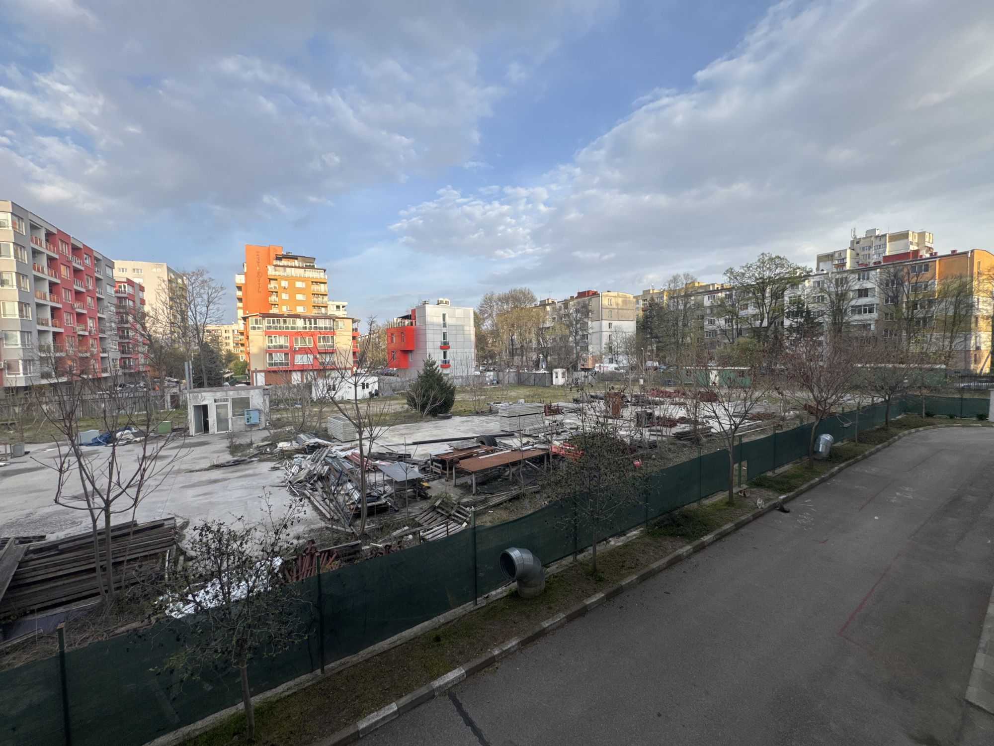 The image shows a vacant lot in Sofia, Bulgaria. The lot is surrounded by buildings, a green fence, and a street. The buildings are a mix of residential and commercial. There is a lot of debris, including wood, metal, and concrete. The lot is overgrown with weeds and grass. There is a sewer pipe in the foreground on the right. The sky is cloudy. There is no visible human activity.