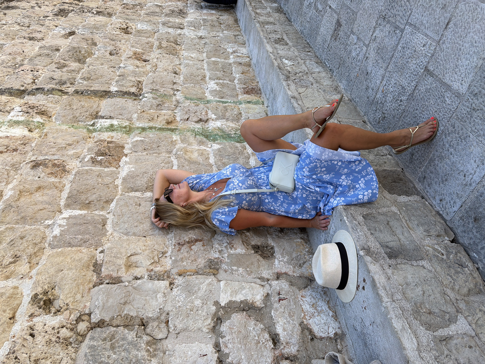 A blonde woman wearing a blue floral dress and gold sandals is sitting on a stone ledge in Dubrovnik, Croatia. She is leaning against a textured stone wall, with her hand resting on her head. A straw hat with a black ribbon lies beside her on the ledge. A white crossbody bag is slung across her body. The ground is paved with large, irregular stone blocks. The scene appears to be outdoors, possibly in the historic Old Town of Dubrovnik.