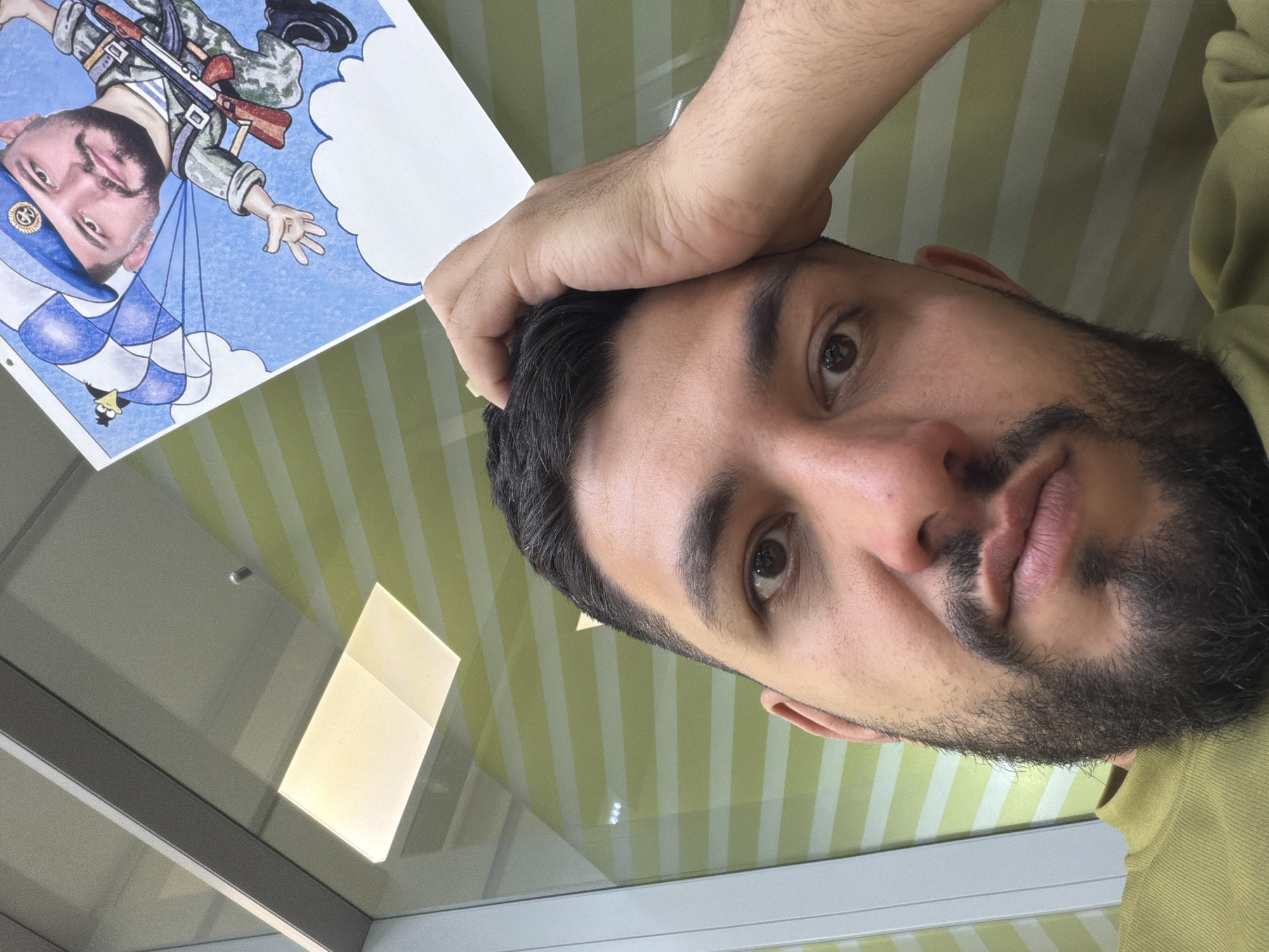 A close-up, rotated image shows a man with dark hair and a beard. He is looking directly at the camera, with his hand near his forehead. To the left of the man's head is a drawing of a character resembling him, wearing a military uniform and a parachute, against a blue sky background. The background consists of green and white striped walls. The location is unidentifiable as Krasnoyarsk, Russia based on the given image.