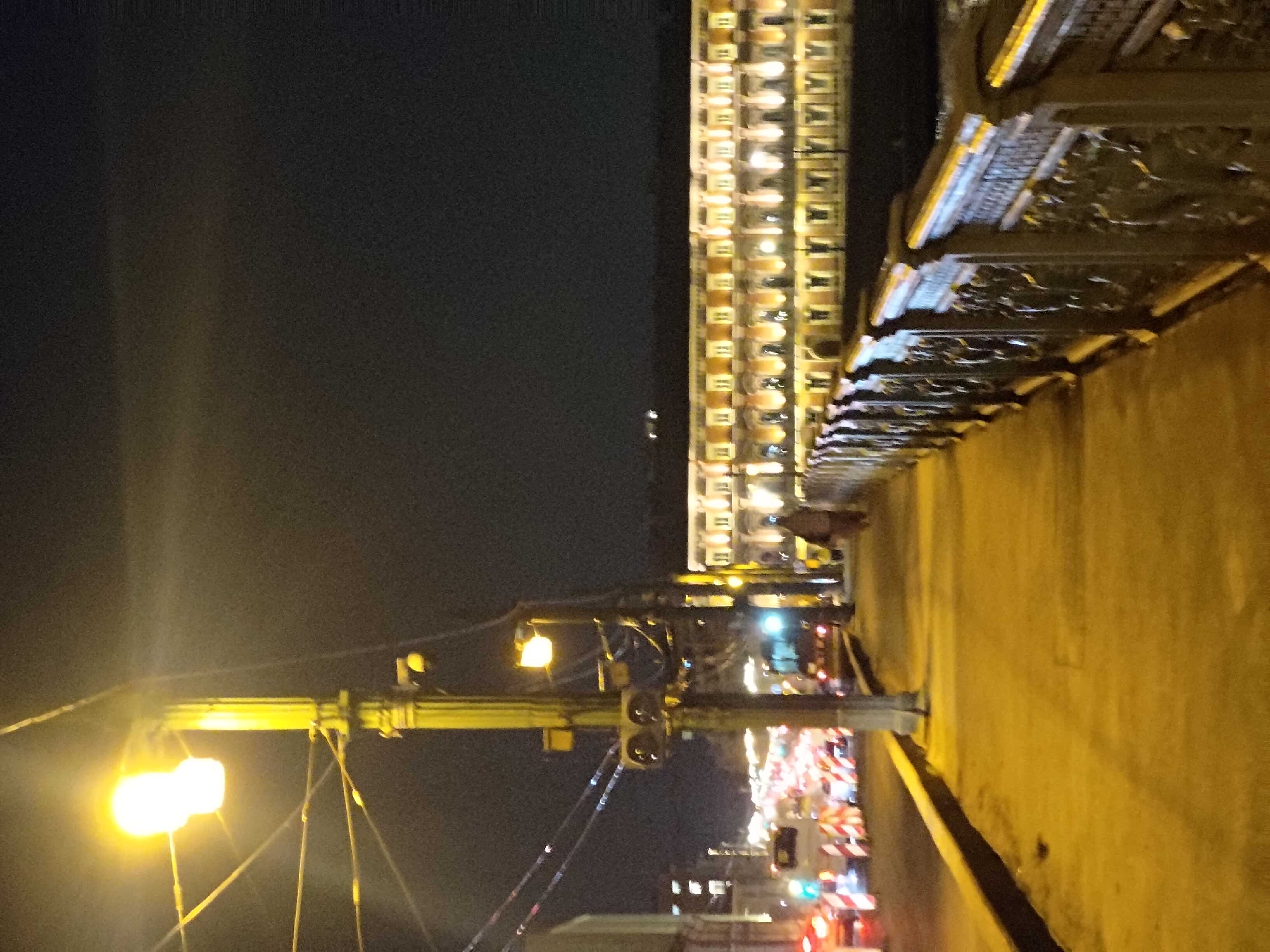 The image depicts a nighttime scene along a city embankment in Saint Petersburg, Russia. A wide, illuminated sidewalk runs along the right side of the frame, bordered by an ornate metal railing. Across a body of dark water to the right, a grand, long building with numerous brightly lit windows stretches across the horizon, its facade reflecting in the water. On the left side of the sidewalk, a street is visible, with traffic lights and a line of vehicles, some with their brake lights illuminated, suggesting a moderate traffic flow. Several streetlights are present, one prominently in the foreground, casting a warm yellow glow. A single pedestrian, wearing a dark, hooded garment, walks away from the camera on the sidewalk, further down. The overall atmosphere is one of urban nocturnal activity under artificial illumination.