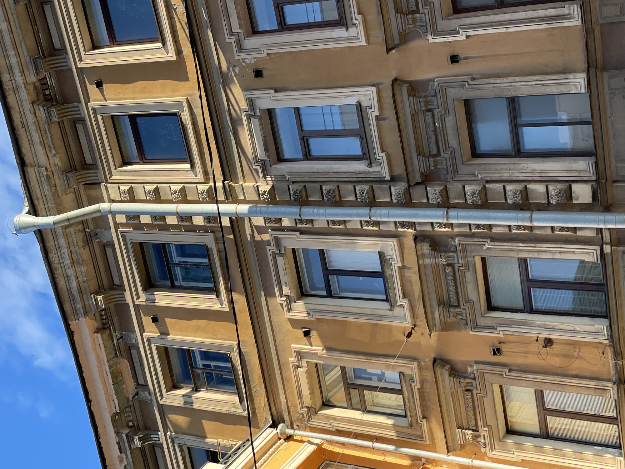 The image presents a low-angle view of the facade of a building in Saint Petersburg, Russia. The building is multi-storied and features a symmetrical arrangement of rectangular windows. The window frames are adorned with decorative architectural elements, including moldings and small reliefs. The exterior of the building is painted in shades of tan and beige, with some sections exhibiting signs of age and weathering, like peeling paint. A metal rain gutter runs horizontally across the facade, obscuring parts of the windows and decorative elements. The sky, a clear blue, is visible in the lower left corner. Electrical wiring is visible along the facade between windows. The perspective is tilted, creating a dynamic angle.