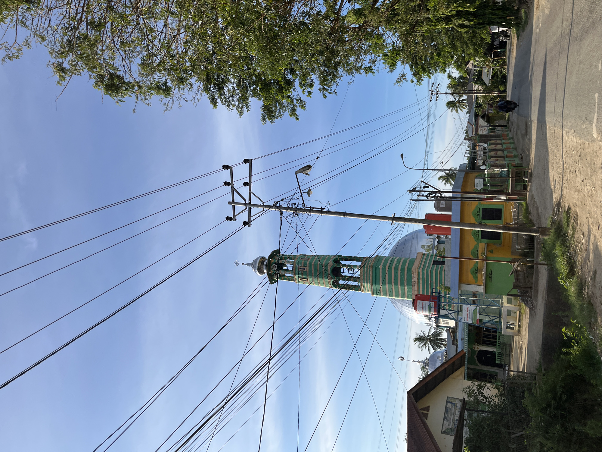 The image captures a street scene in Alurcucur, Indonesia, under a bright blue sky with wispy clouds. Overhead, a dense network of power lines crisscrosses the frame, supported by utility poles. To the left, a mosque with a distinctive green and white striped minaret and a large dome is visible. Adjacent to the mosque are several colorful buildings, some with painted facades. A water tank with "PERTAMINA PEDULI" branding stands on a metal structure near the mosque. A dirt road runs alongside the buildings, leading to an asphalt road in the foreground. A person is seen riding a motorcycle on the asphalt road, heading away from the camera. Lush green trees line the right side of the road, casting shadows on the scene. The overall impression is of a sunlit, modest urban or semi-urban environment.